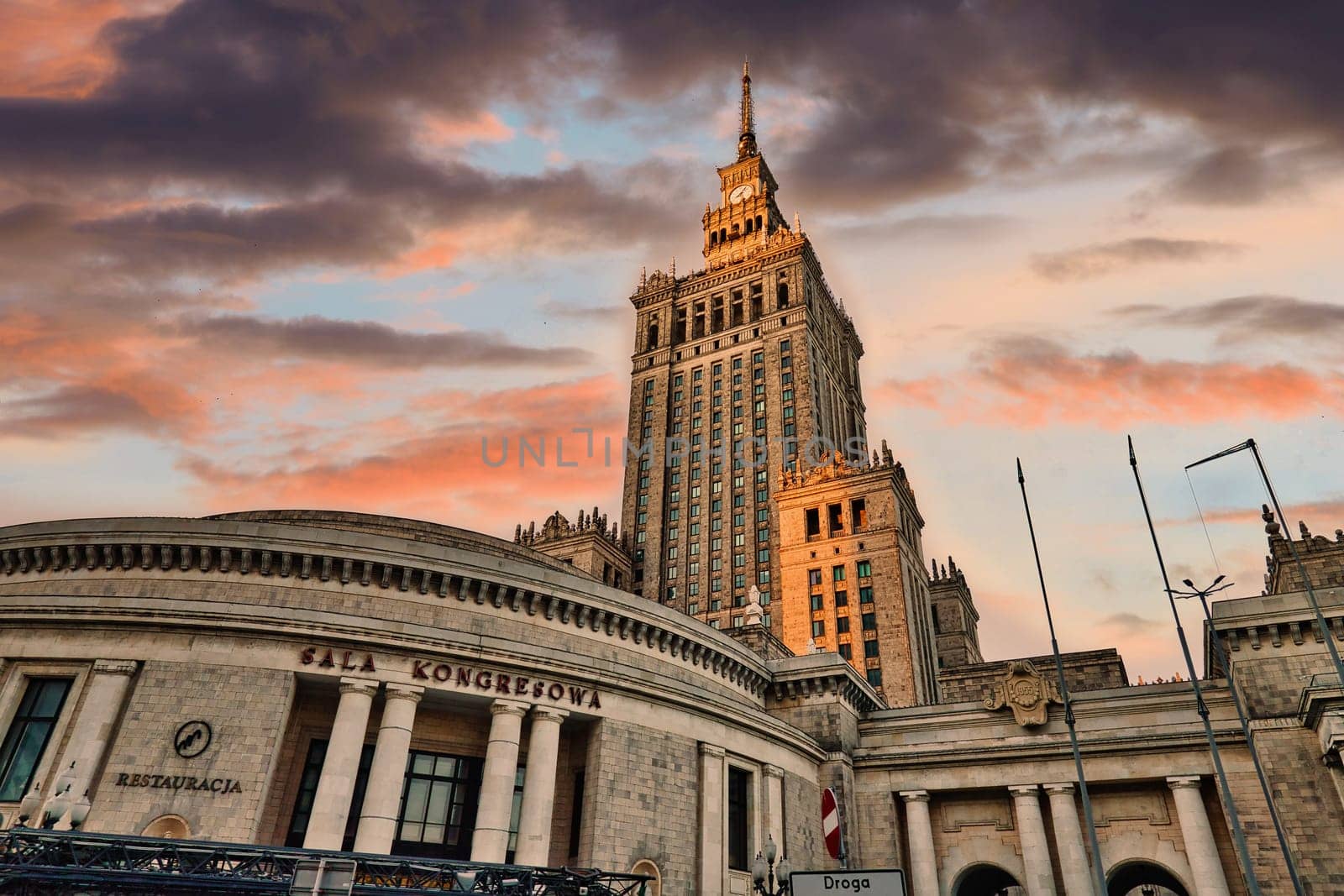 A striking image of the Palace of Culture and Science in Warsaw, Poland, captured at sunset. The towering structure, a symbol of post-war architecture, dominates the skyline with its impressive design and intricate details. The colorful sky adds drama and contrast, making the iconic building stand out even more. Perfect for themes related to travel, history, and urban architecture.