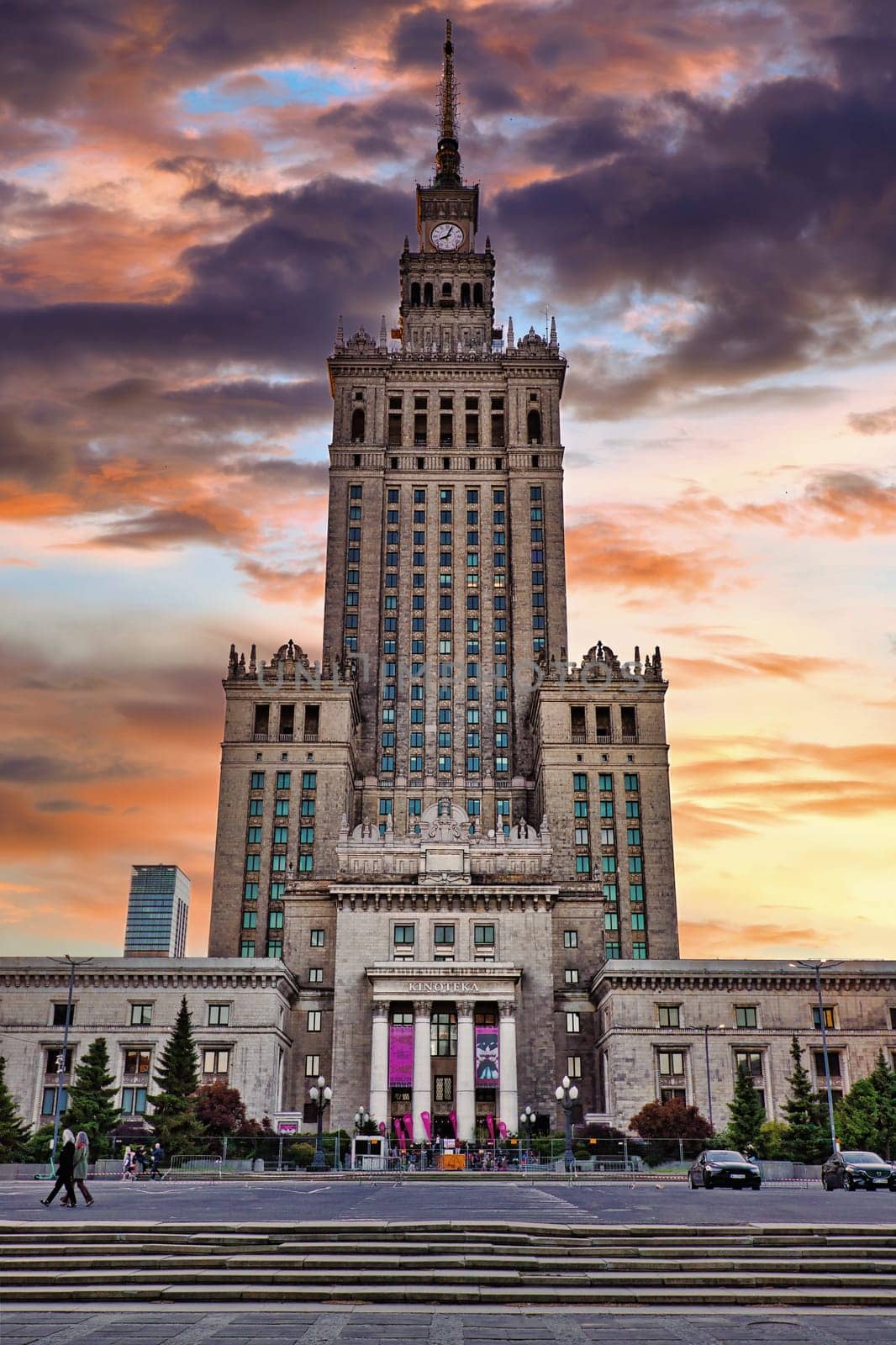 A majestic front view of the Palace of Culture and Science in Warsaw, Poland, taken during sunset. The building, an iconic symbol of Warsaw, stands tall with its grand architecture and historical significance, built in the 1950s as a gift from the Soviet Union. The vivid colors of the sky create a stunning contrast against the stone facade, making it a striking image of one of Warsaw most famous landmarks. Ideal for themes related to architecture, history, and travel.