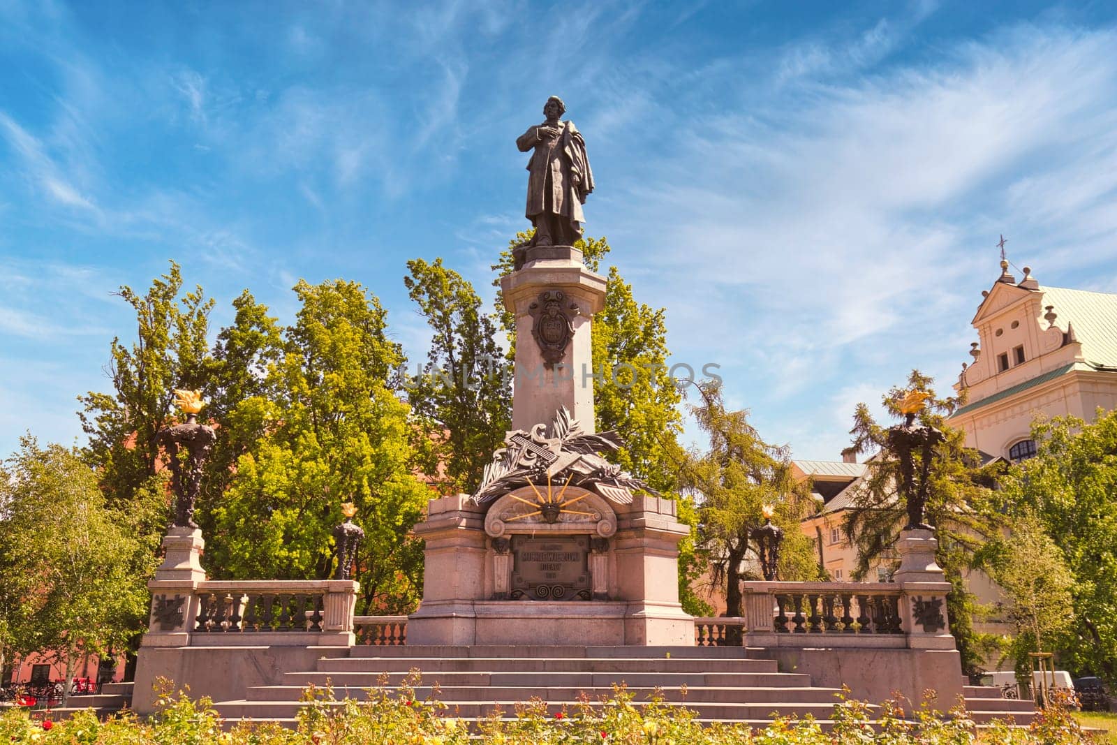 A grand view of the Adam Mickiewicz Monument located in Warsaw, Poland, set against a bright blue sky. This statue commemorates one of Poland greatest poets and is a significant cultural and historical landmark. Surrounded by lush greenery and intricate sculptures, the monument stands proudly in a public square, attracting visitors and locals alike. Ideal for themes related to history, culture, and travel.
