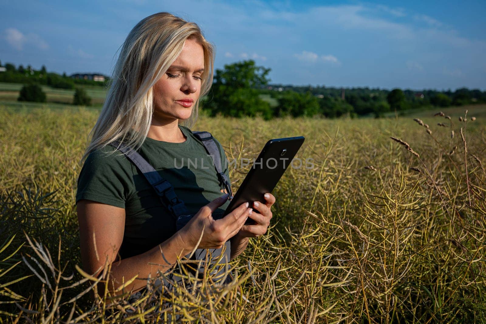 A woman farmer is checking the quality of rapeseed on the middle of the field and holding a tablet. She is checking the information about her harvest and she seems worried about the quality of rapeseed. She owns a rapeseed field and is checking this year's harvest. She is an experienced farmer, caring for her crops in a vast field. She has blonde hair. The soil is fertile and productive. The rapeseed is yellow and ready for harvest. Satisfied with the bountiful and lavish harvest, the woman farmer, adorned in working clothes and with tanned skin, gazes upon the expansive yellow landscape with a sense of fulfillment. With the blue sky adorned with fluffy white clouds overhead, she stands in the middle of the field, her spirit merging with the warmth of the sun, celebrating the culmination of her dedication and the richness of the land's yield.