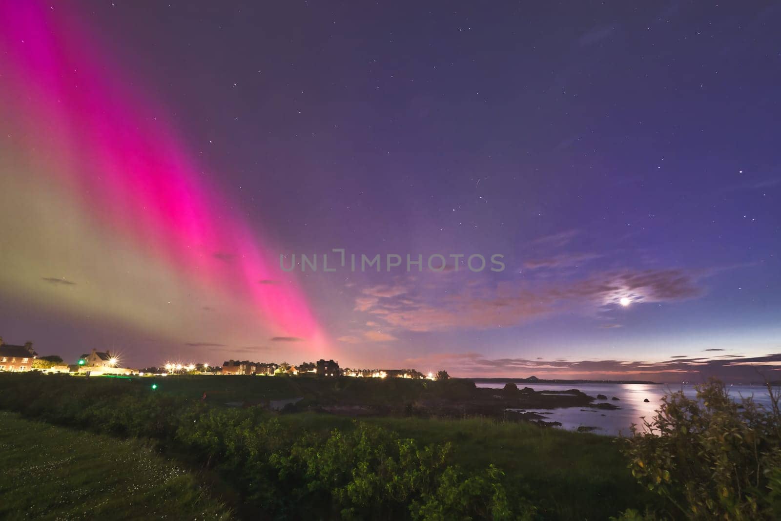 A stunning capture of the Aurora Borealis lighting up the sky over North Berwick coastline. The striking pink and green auroras contrast beautifully with the darkening sky and calm coastal waters, creating a serene yet dramatic scene. The town lights in the distance, along with the crescent moon, add to the ethereal beauty of this rare phenomenon. Perfect for use in travel, nature, and astrophotography themes.