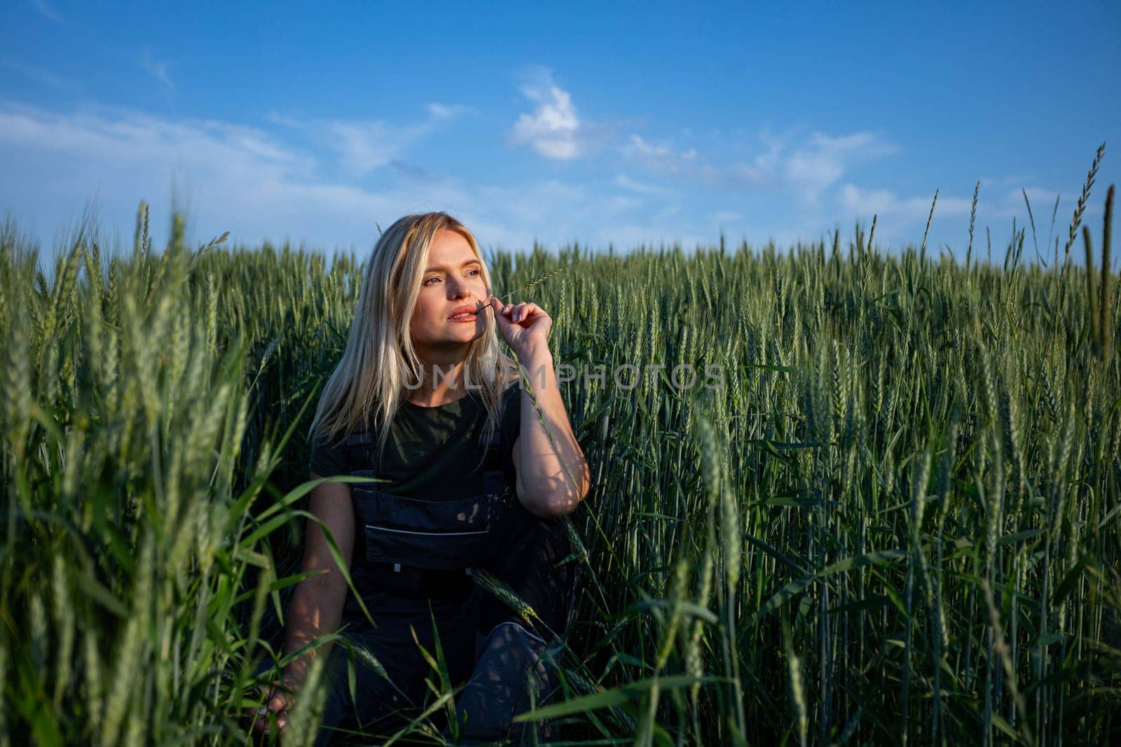 In the tranquil expanse of a rustic wheat field, a female farmer squats, holding a wheat stalk in one hand, contemplatively placing it near her lips. With sun-kissed skin and clad in work attire, she embodies the essence of rural life. Her expression is one of introspection, leaving viewers to ponder whether she is content with the harvest or harbors concerns. She is looking at the horizon. The vibrant green hue of the wheat stalk contrasts against the golden tones of the field, adding depth to the scene. Against the backdrop of the sprawling countryside, her presence evokes a sense of agricultural tradition and connection to the land. This image captures the quiet moments of farming, where contemplation mingles with the labor of the harvest. As she gazes thoughtfully upon the wheat, her blonde hair softly frames her face, enhancing the rustic charm of the scene