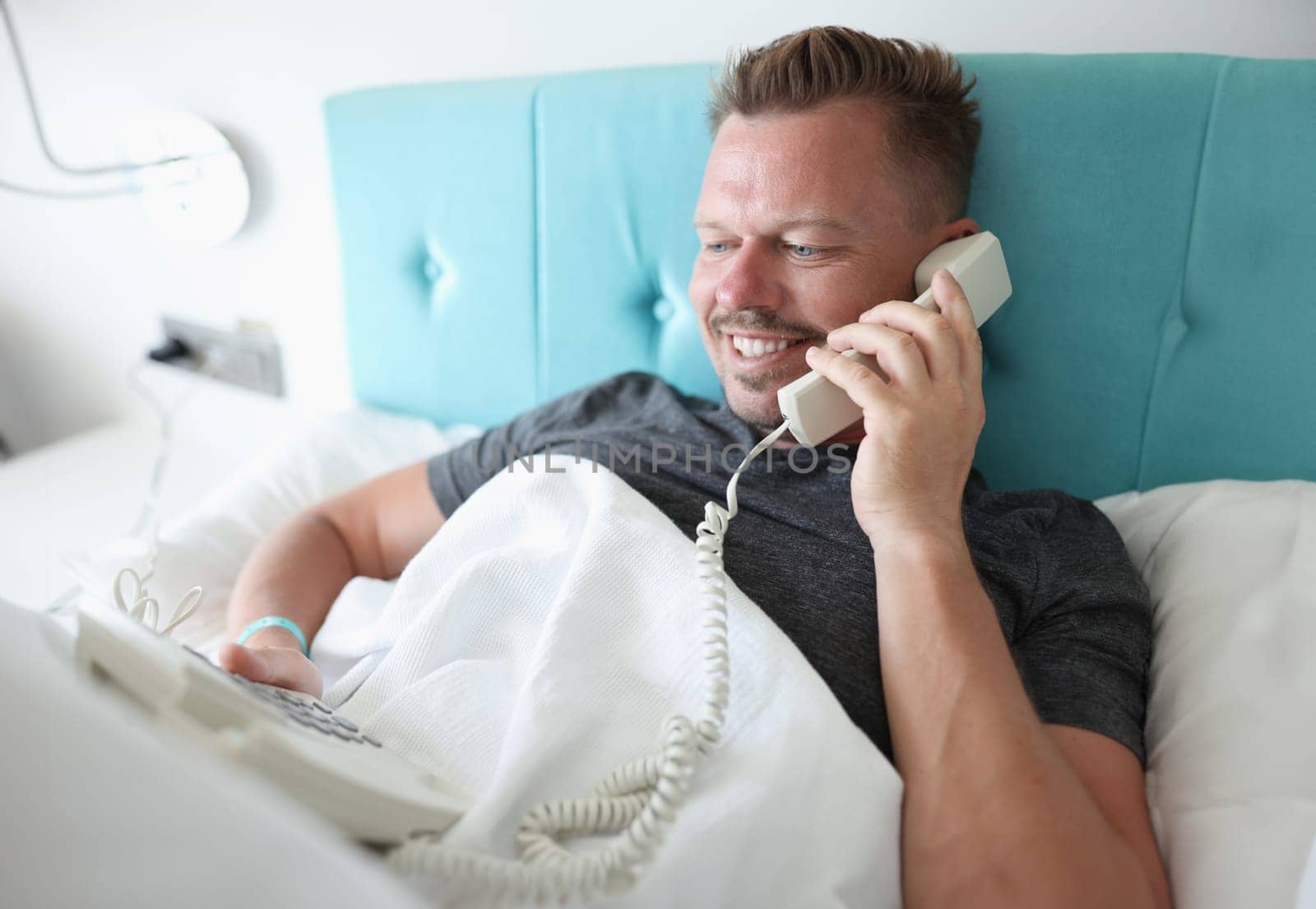 Smiling man lies in bed and talks on phone. Calling administrator to hotel room concept