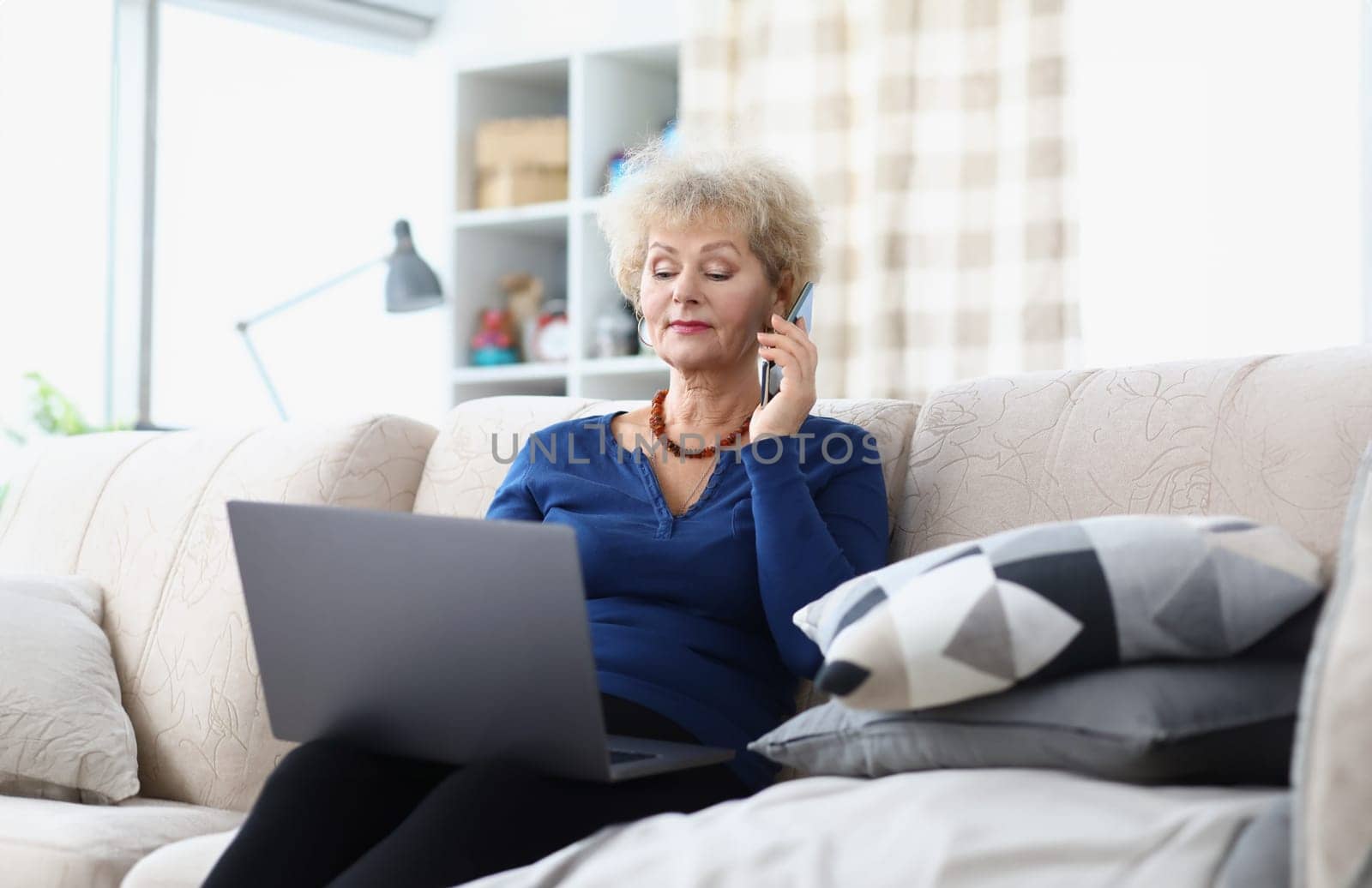 An elderly woman is sitting on couch with laptop and smartphone. Teaching retirees to work remotely concept