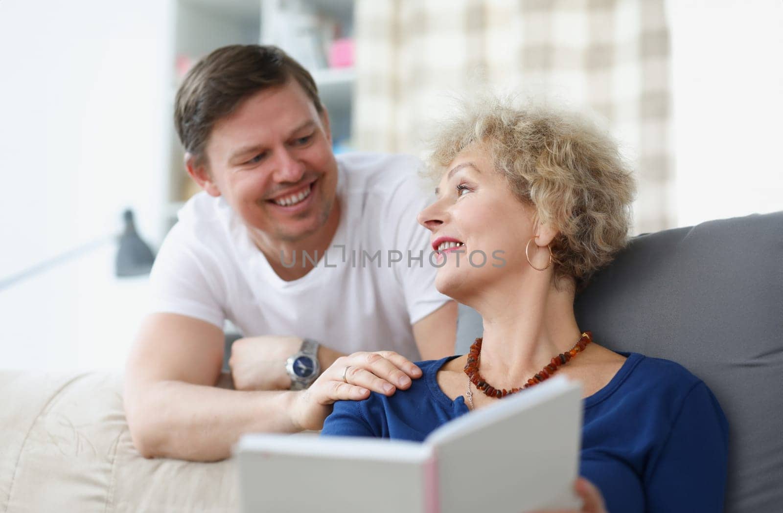 An elderly woman is sitting on sofa and smiling at young man. Relationship between adult children and parents concept