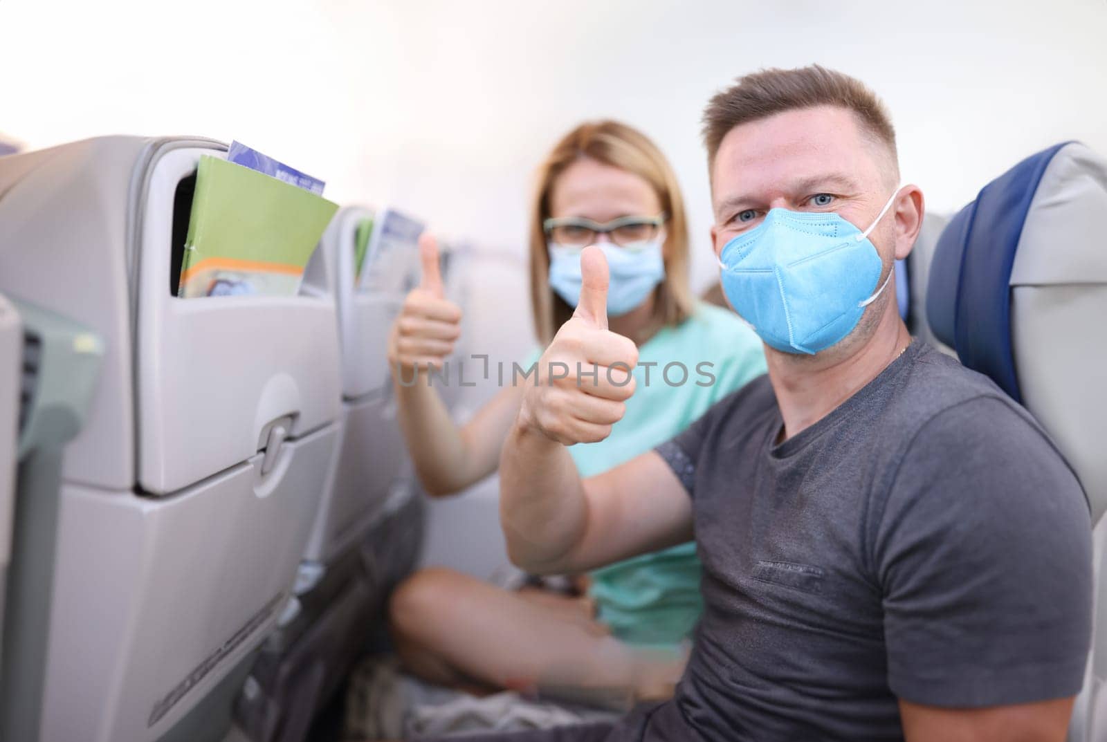 Man and woman in medical protective masks in aircraft cabin hold their thumbs up. Safe flights to coronavirus pandemic concept