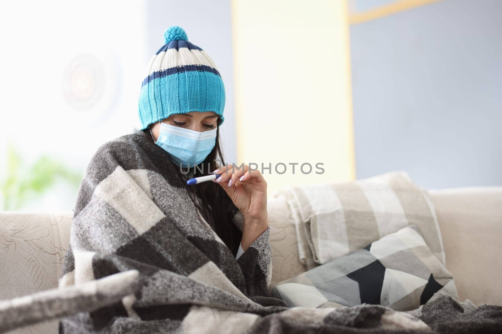 Frozen woman in hat and blankets in protective mask sits on sofa with thermometer. Seasonal diseases and coronavirus pandemic concept