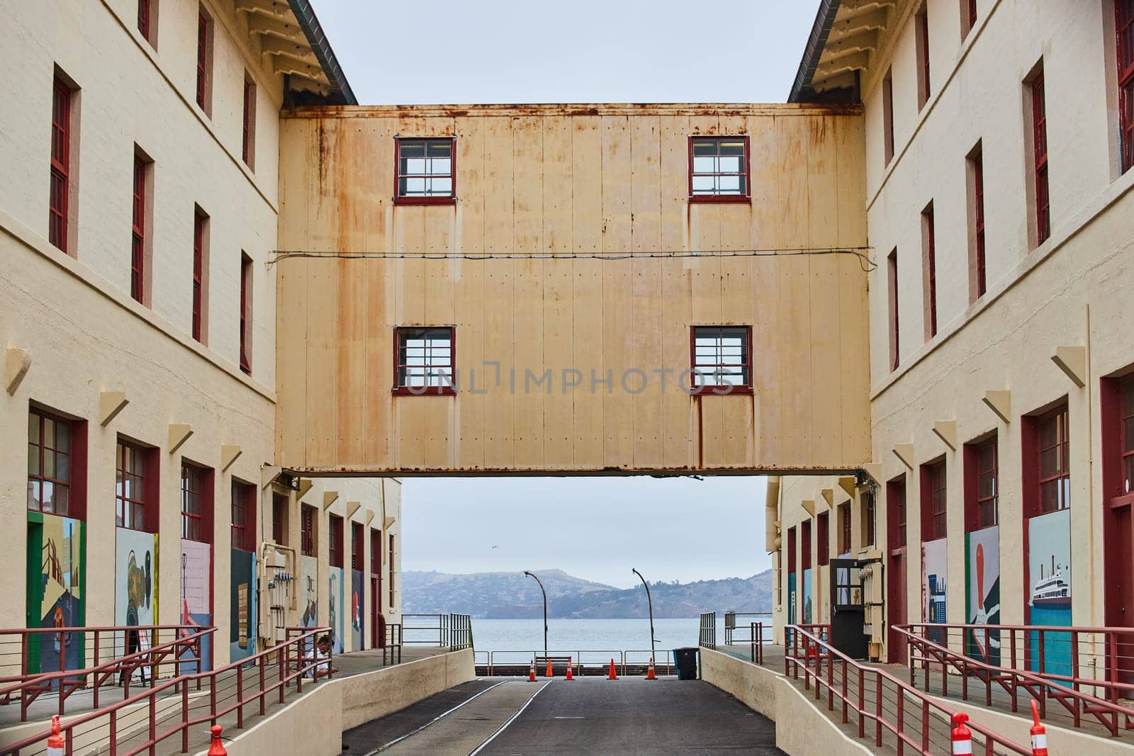 Image of Rusty bridge between two warehouse buildings with road underneath and view of San Francisco Bay