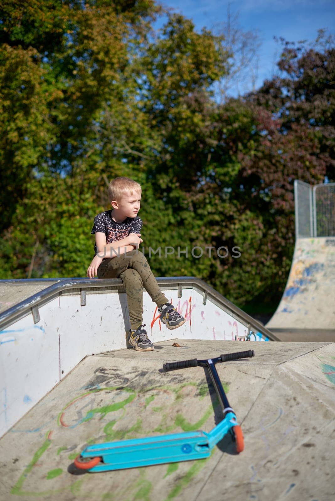 Boy on scooter makes a trick and enjoying his riding in the skate park at cloudy spring day. Young man doing trick on the kick scooter in the park. Full body little boy on the kick scooter on sunny autumn day in skate park. From below of active teen boy with kick scooter standing on ramp in skate park while preparing for performing trick in sunny spring day. by Andrii_Ko