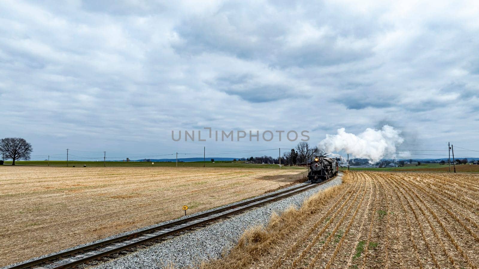 A historic steam train chugs along the tracks, billowing white smoke as it cuts through open fields under a dramatic sky filled with clouds.