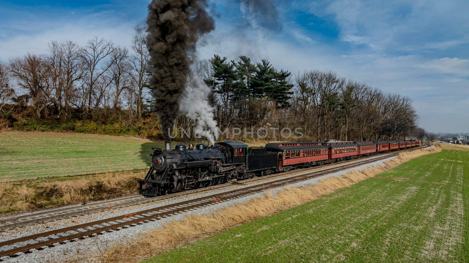 A vintage steam locomotive puffs black smoke as it chugs along railway tracks, bordered by lush green fields and trees under a clear blue sky. Passengers enjoy the scenic ride.