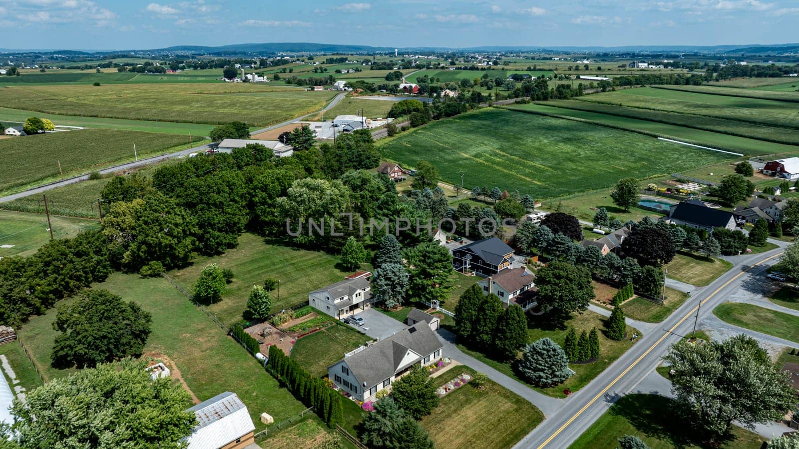 The landscape showcases a sprawling countryside with green fields, dotting houses, and trees under a clear blue sky, illustrating the serene charm of rural living.