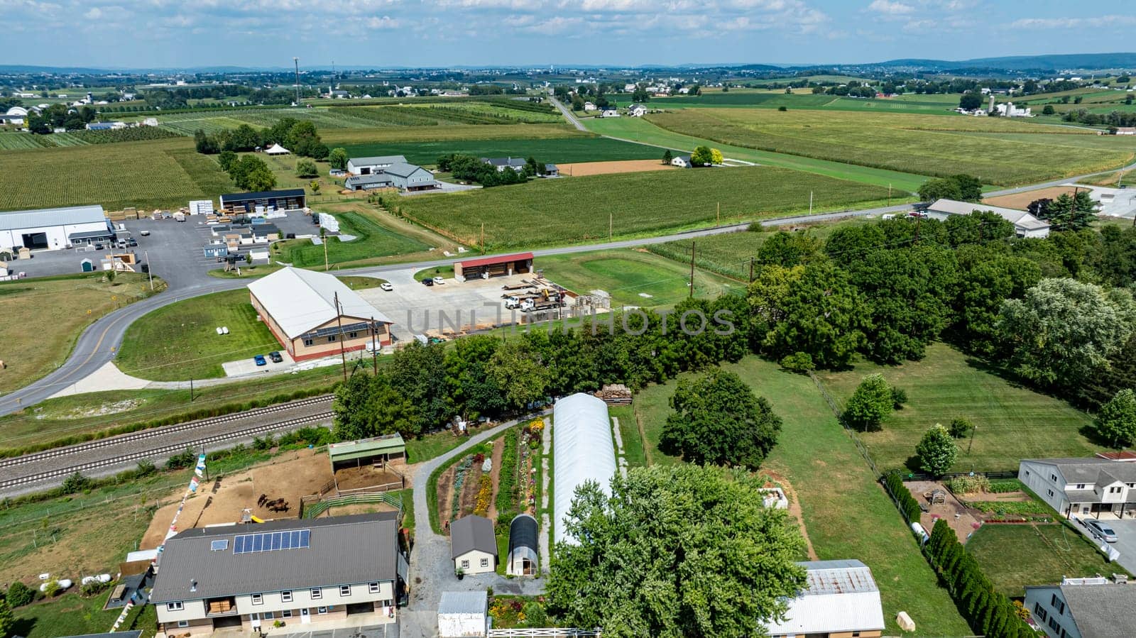 This aerial perspective captures a vast rural landscape featuring multiple farms, green fields, and winding roads under a clear blue sky. The scene highlights the serenity of countryside life.