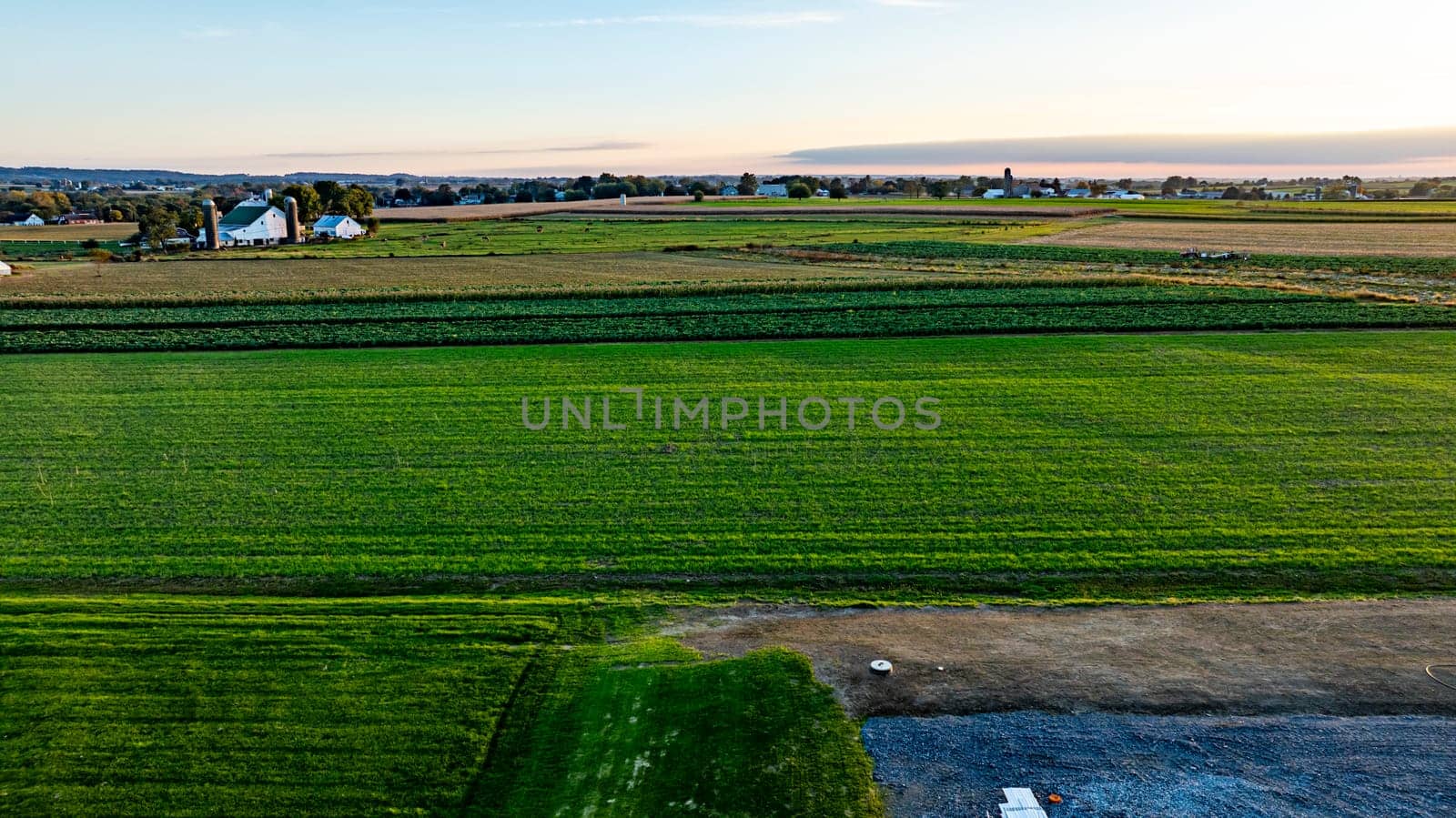 Lush green fields stretch across the landscape as the sun sets, casting a warm glow over distant houses and the serene rural area, inviting a sense of peace and tranquility.