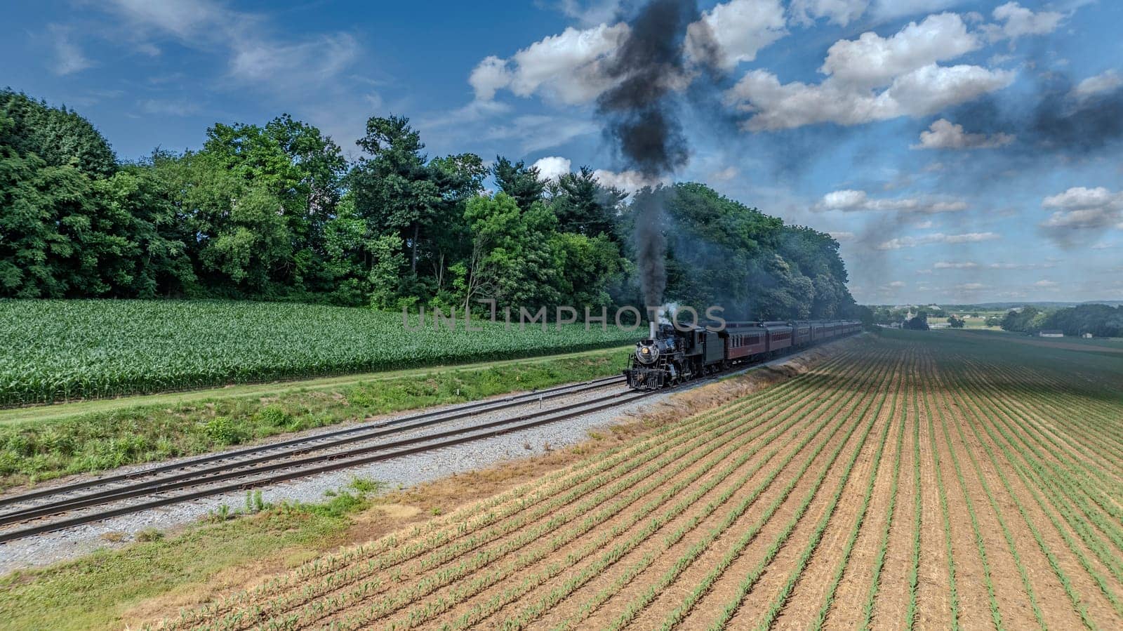 A Black Steam Locomotive Billowing Thick Smoke Pulls Red Passenger Cars Through Lush Green Fields And Forested Areas, Under A Clear Blue Sky With Puffy Clouds, Creating A Picturesque Countryside Journey.