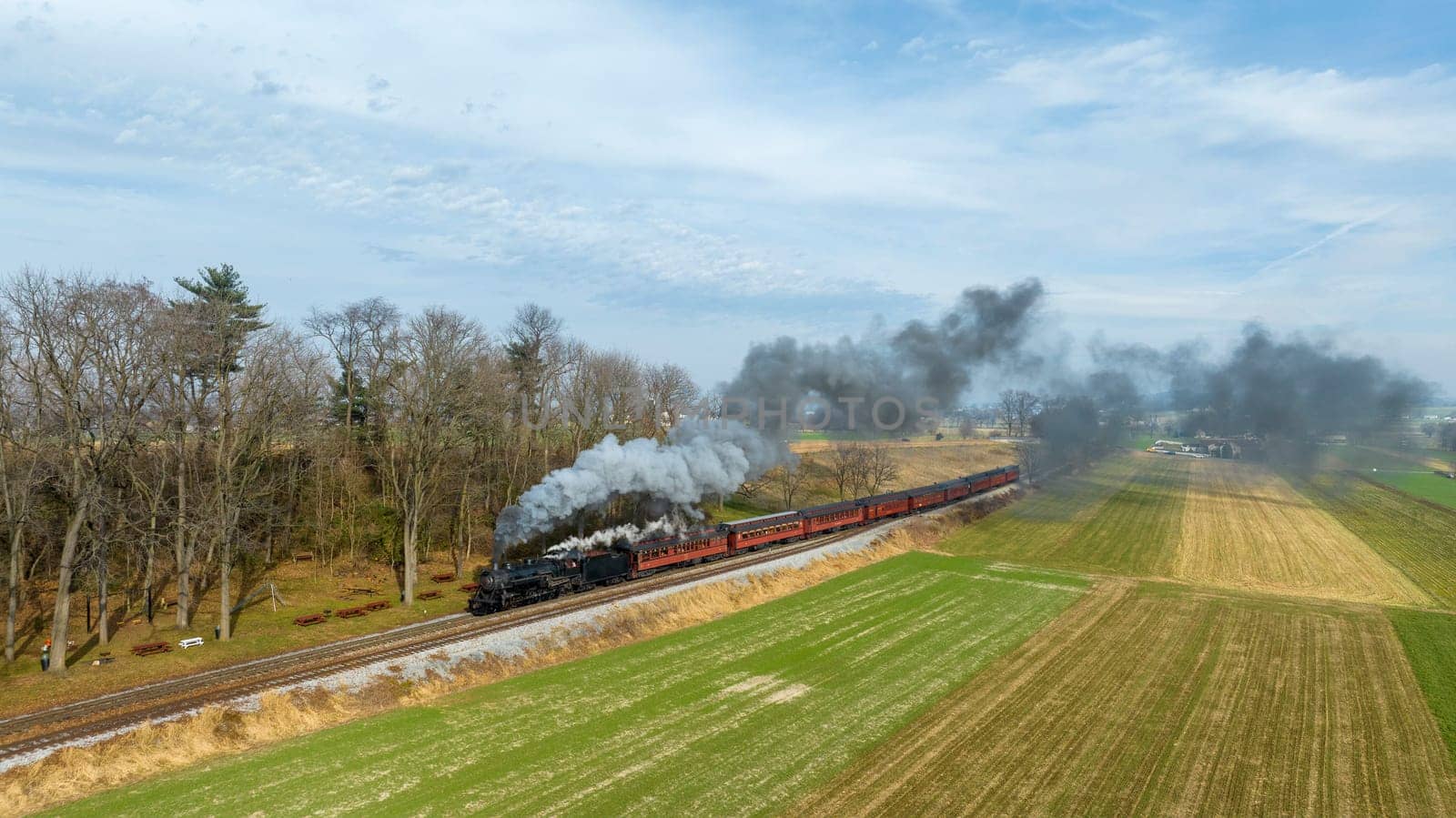A steam locomotive pulls a vintage train through a picturesque countryside, emitting smoke and steam against a backdrop of fields and trees under a bright sky.