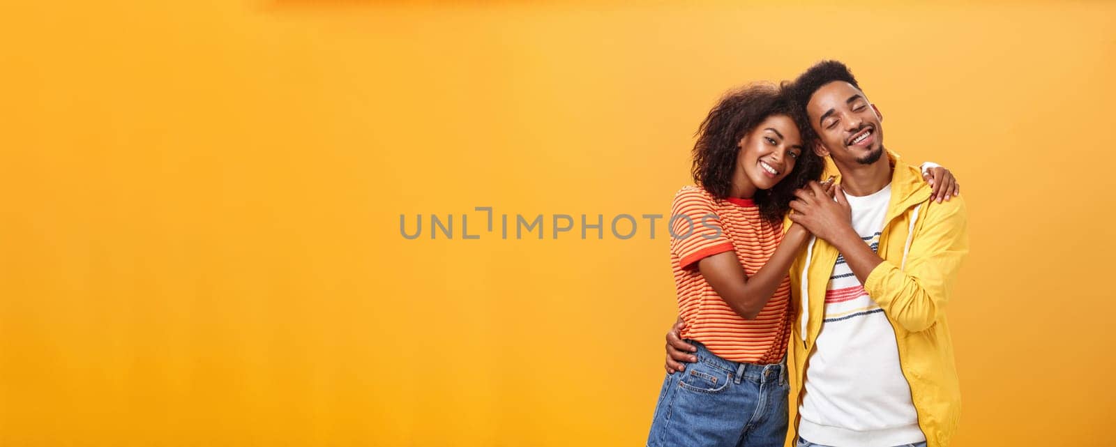 They love each other. Portrait of two charming african american man and woman in relationship hugging with heartwarming smile touching hands smiling gently posing against orange background.