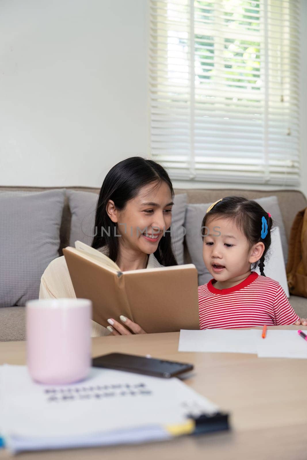 Asian mother and young daughter engaged in reading a book together, creating a warm and educational atmosphere in a cozy living room.