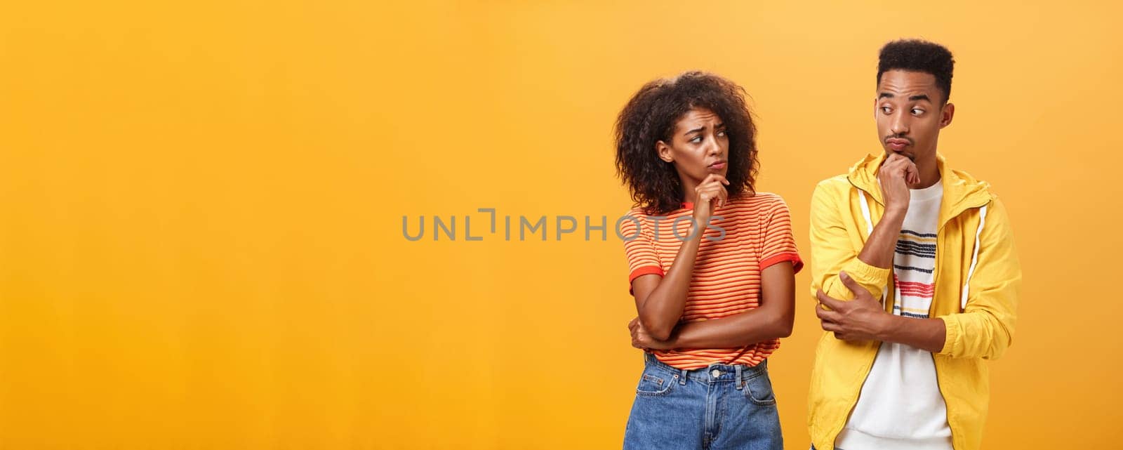 Silly confused girl standing gloomy and worried pursing lips and frowning holding hand on chin looking at boyfriend confused while guy glancing at girlfriend with curious expression over orange wall. Lifestyle.