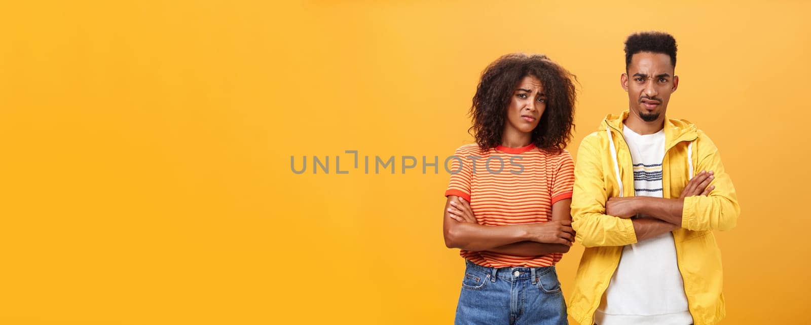 Two friends dislike lame idea of mate. Portrait of dissatisfied unimpressed african american man and woman crossing arms on chest in aversion frowning doubtful and disappointed over orange wall.