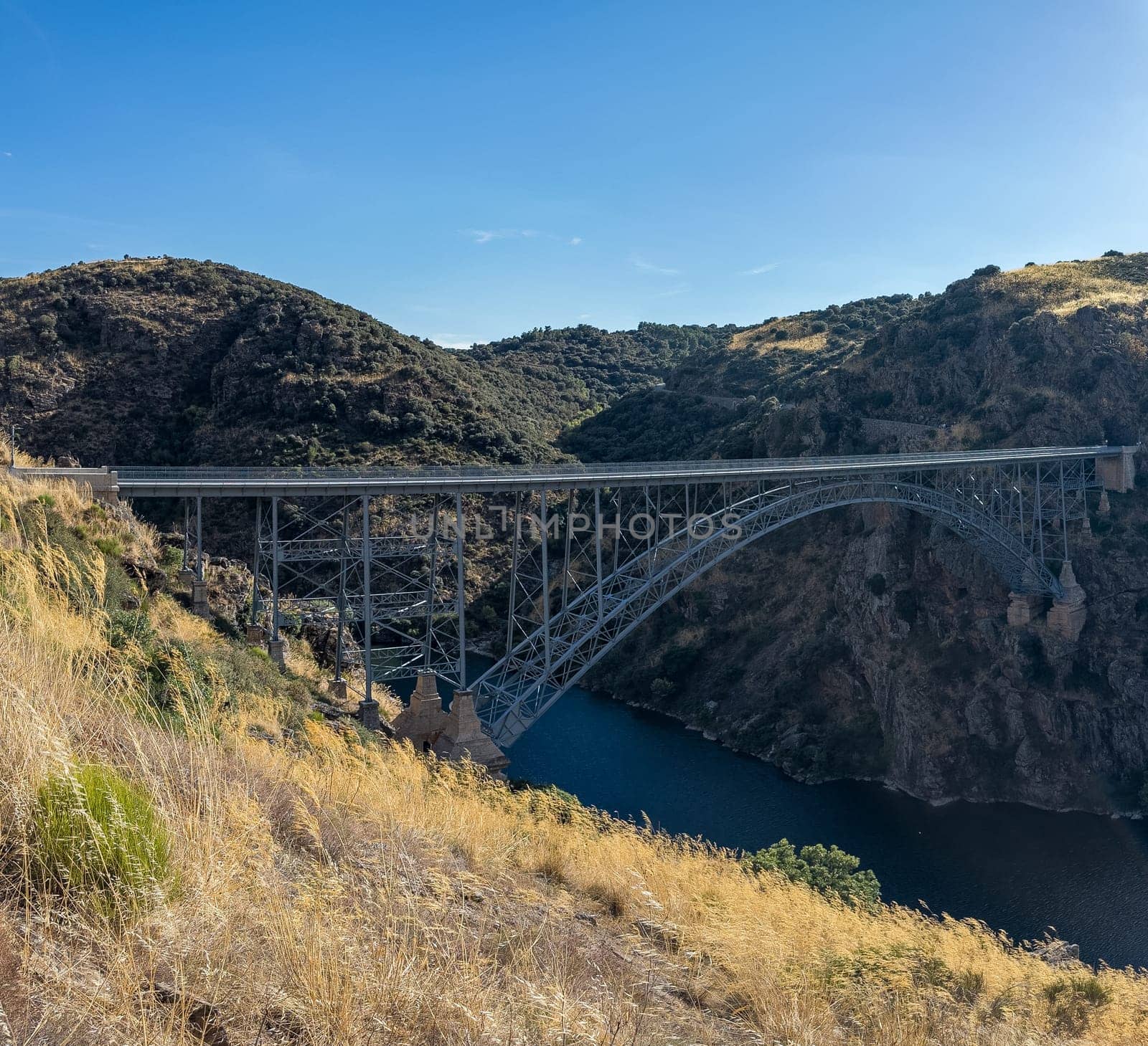 A steel arch bridge crosses a tranquil river, surrounded by rugged hills beneath a clear sky.