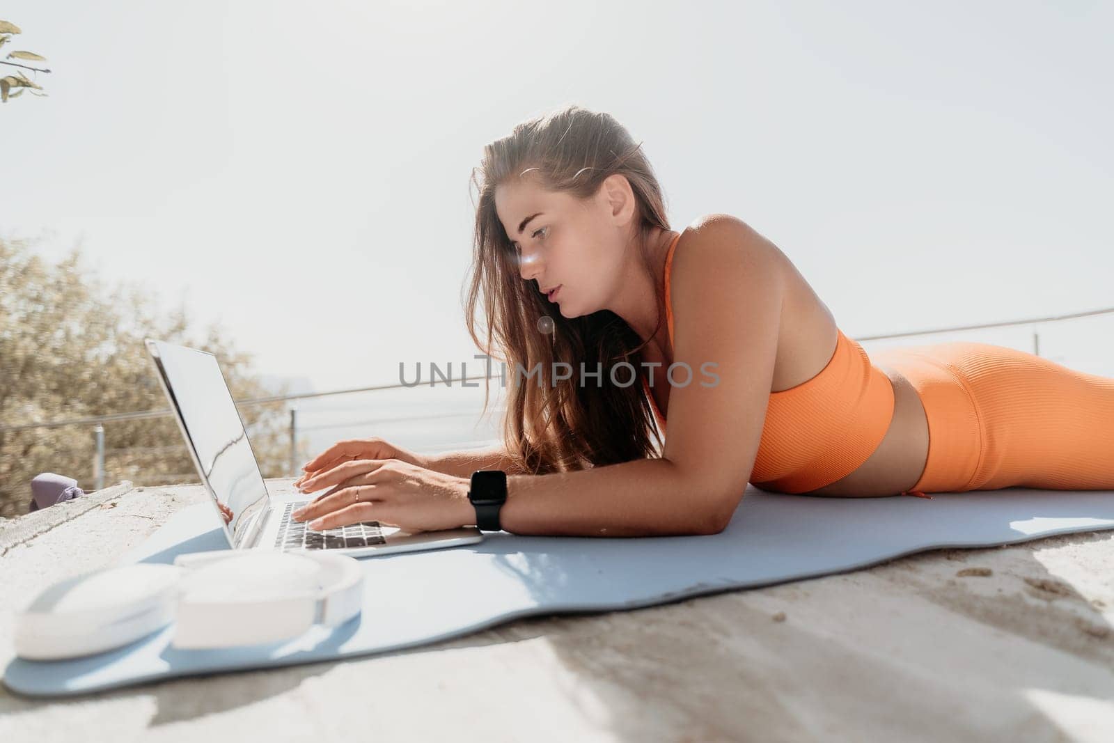 A woman wearing an orange sports bra and leggings lies on a yoga mat while working on a laptop in a sunny outdoor setting.