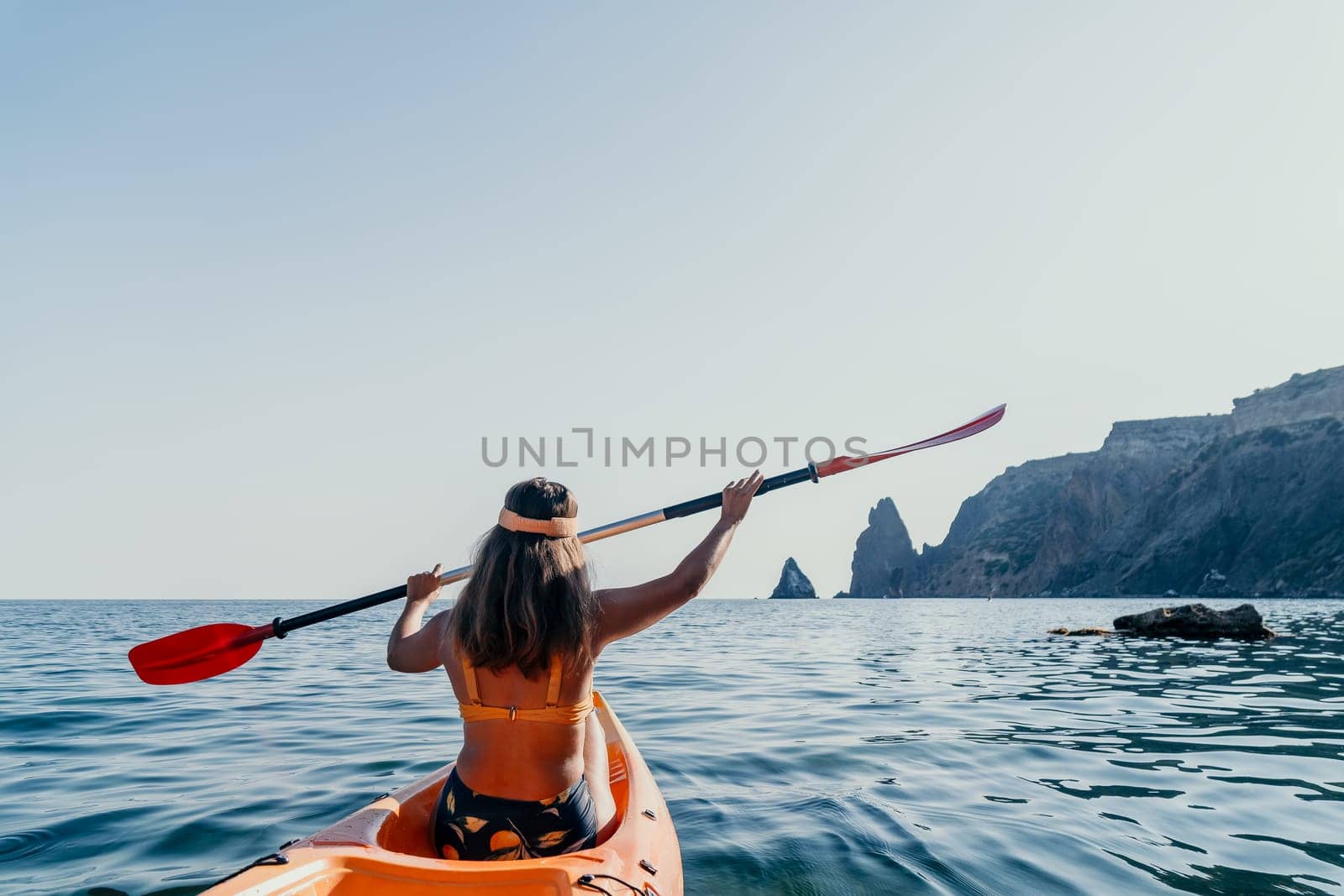 A woman paddles a kayak towards a rocky cliff in the ocean. The sun shines brightly and the water is calm.
