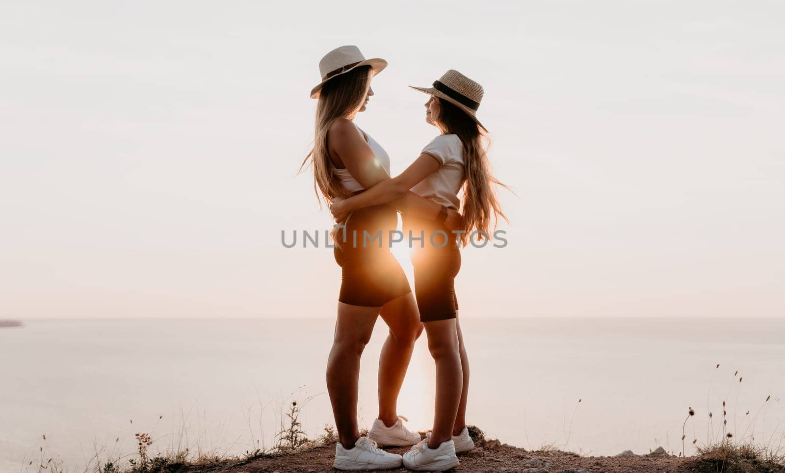 Two women in hats stand on a cliff overlooking the ocean, embracing as the sun sets behind them.