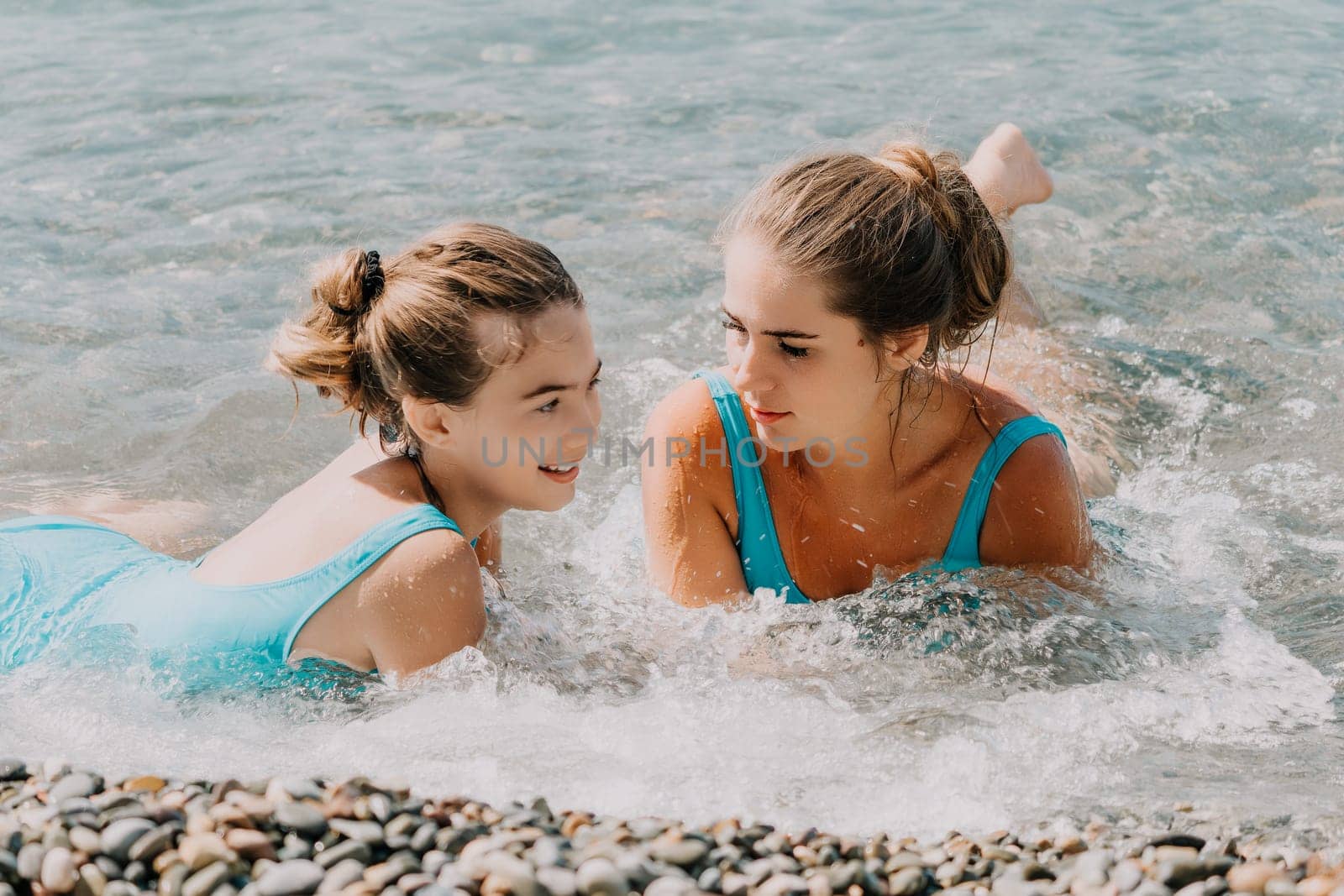 Two young women in blue swimsuits are lying in the shallows of the ocean, one looking at the camera and the other looking at her companion, their hair is wet and they are covered in water.