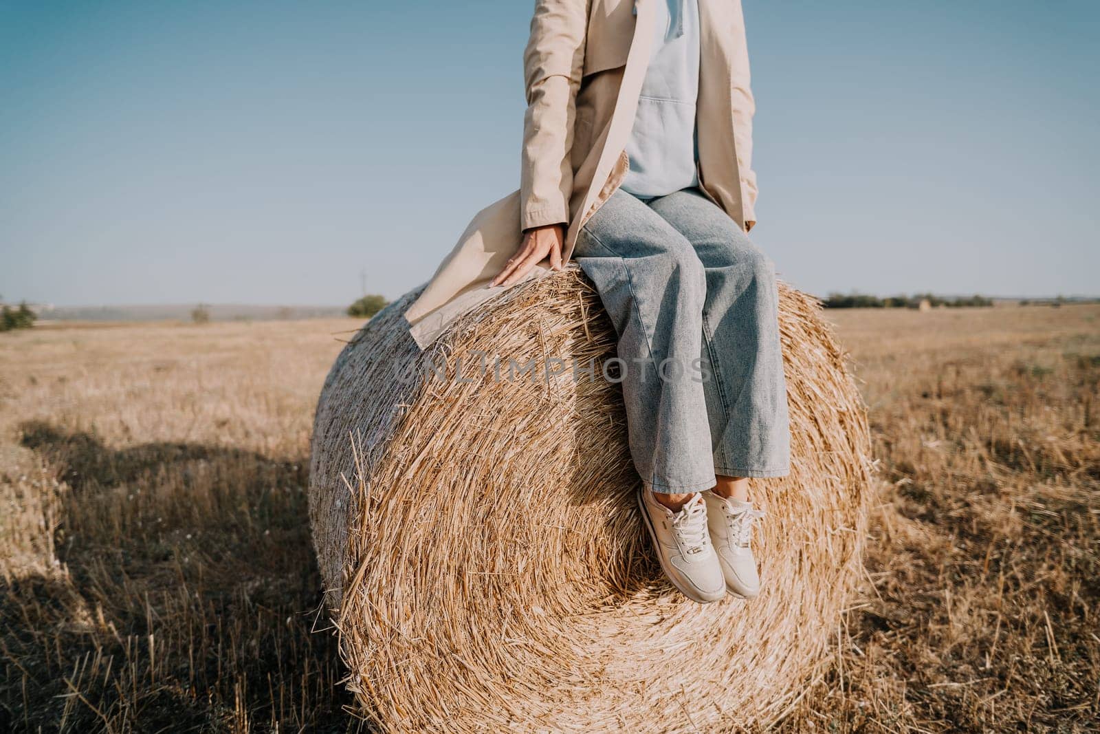 A woman wearing a trench coat and blue jeans sits on a large hay bale in a field. The sky is blue and the field is brown.