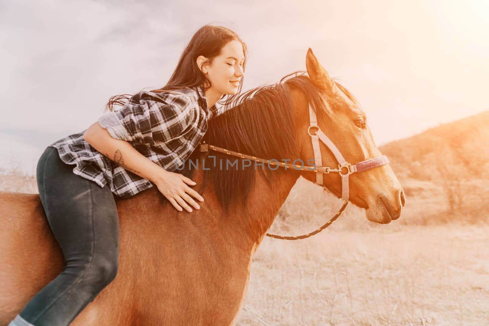 A young woman in a plaid shirt rides a brown horse with a sunset in the background.