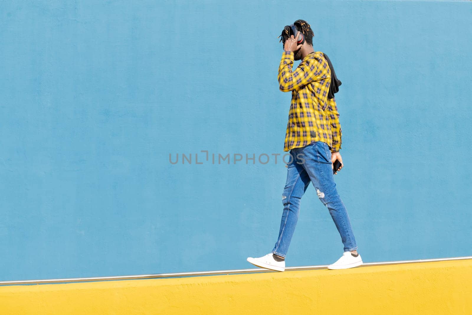 unrecognizable young black man with headphones walking along a yellow wall and a blue wall in the background with his smartphone in his hand, technology and lifestyle concept, copy space for text