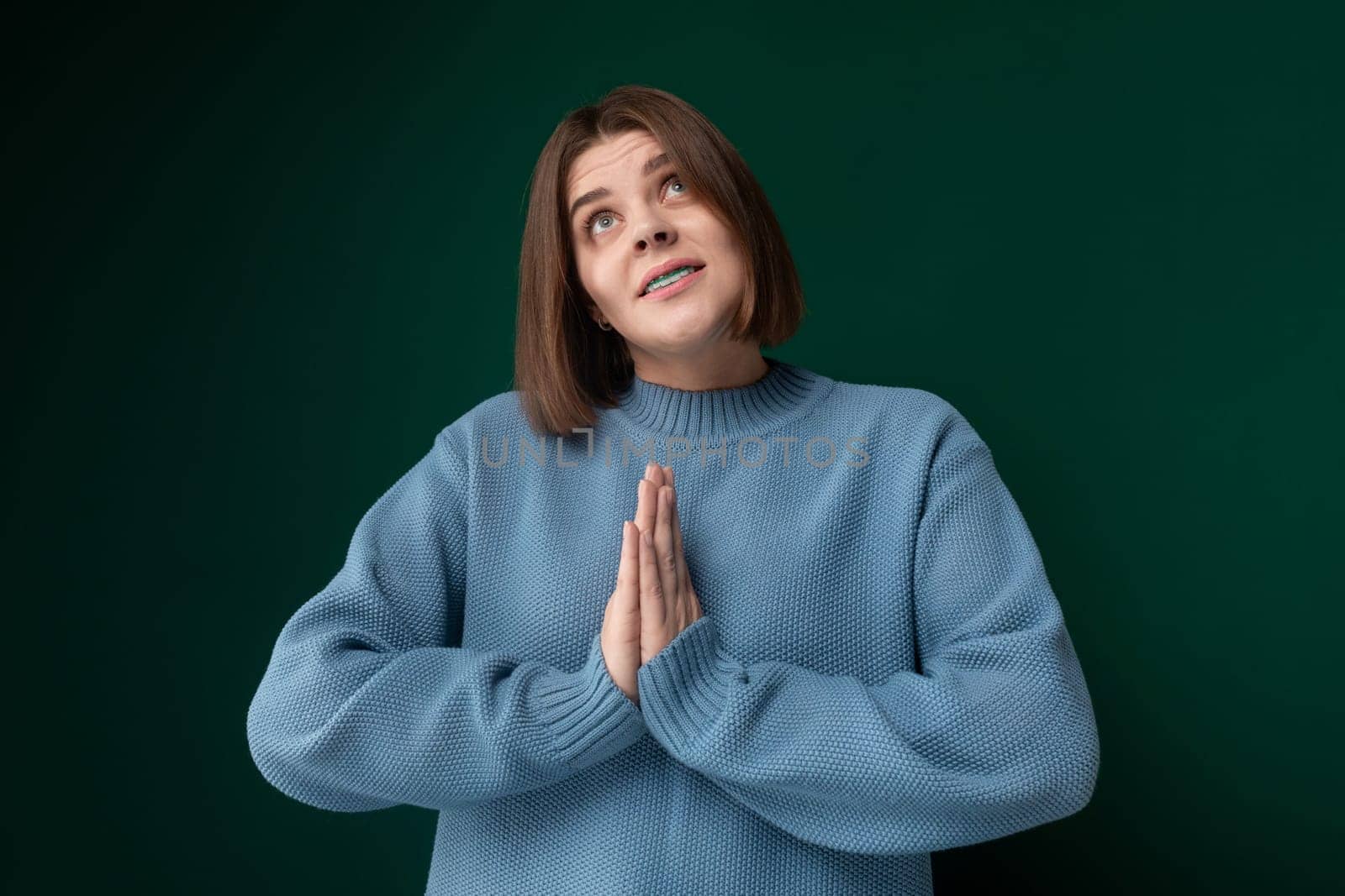 A woman wearing a blue sweater is shown in a prayerful pose, with her hands clasped together and eyes closed. She appears to be deep in prayer, with a solemn expression on her face.