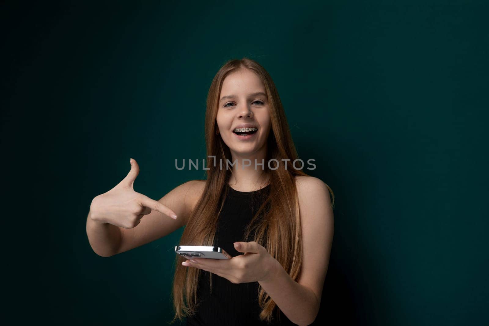 A young girl is holding a cell phone in one hand, looking directly at the screen, and giving a thumbs up with her other hand. She seems happy and enthusiastic about something on the phone.