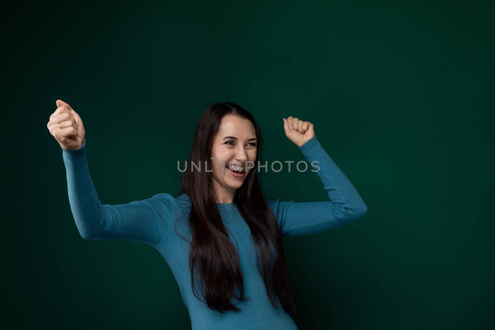 A woman wearing a blue shirt is joyfully lifting her arms upwards in celebration or victory. Her arms are extended fully, and her face displays a sense of triumph or happiness.