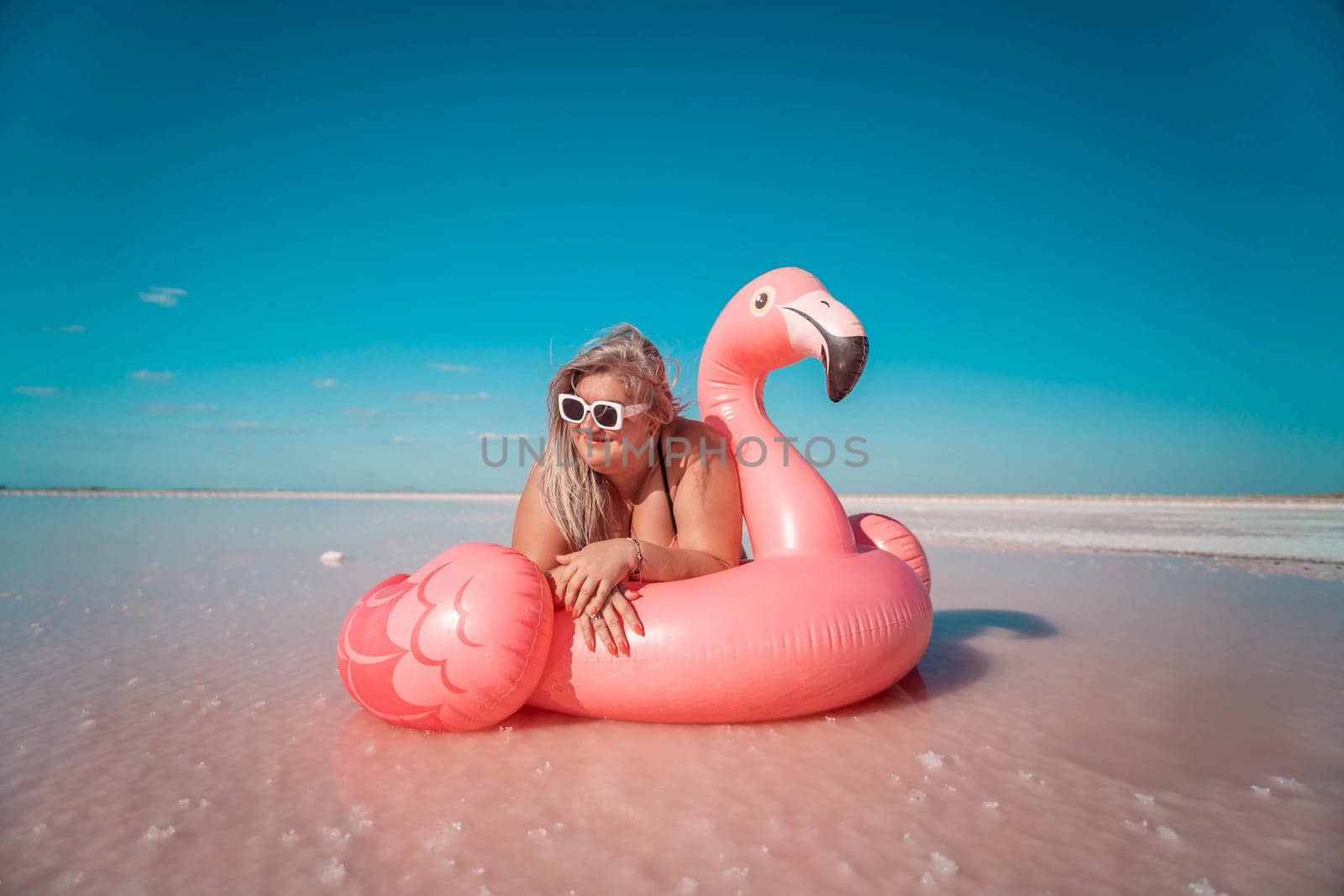 A woman in sunglasses lays on a pink flamingo inflatable in a shallow body of water with a pink tint. The sky is blue.