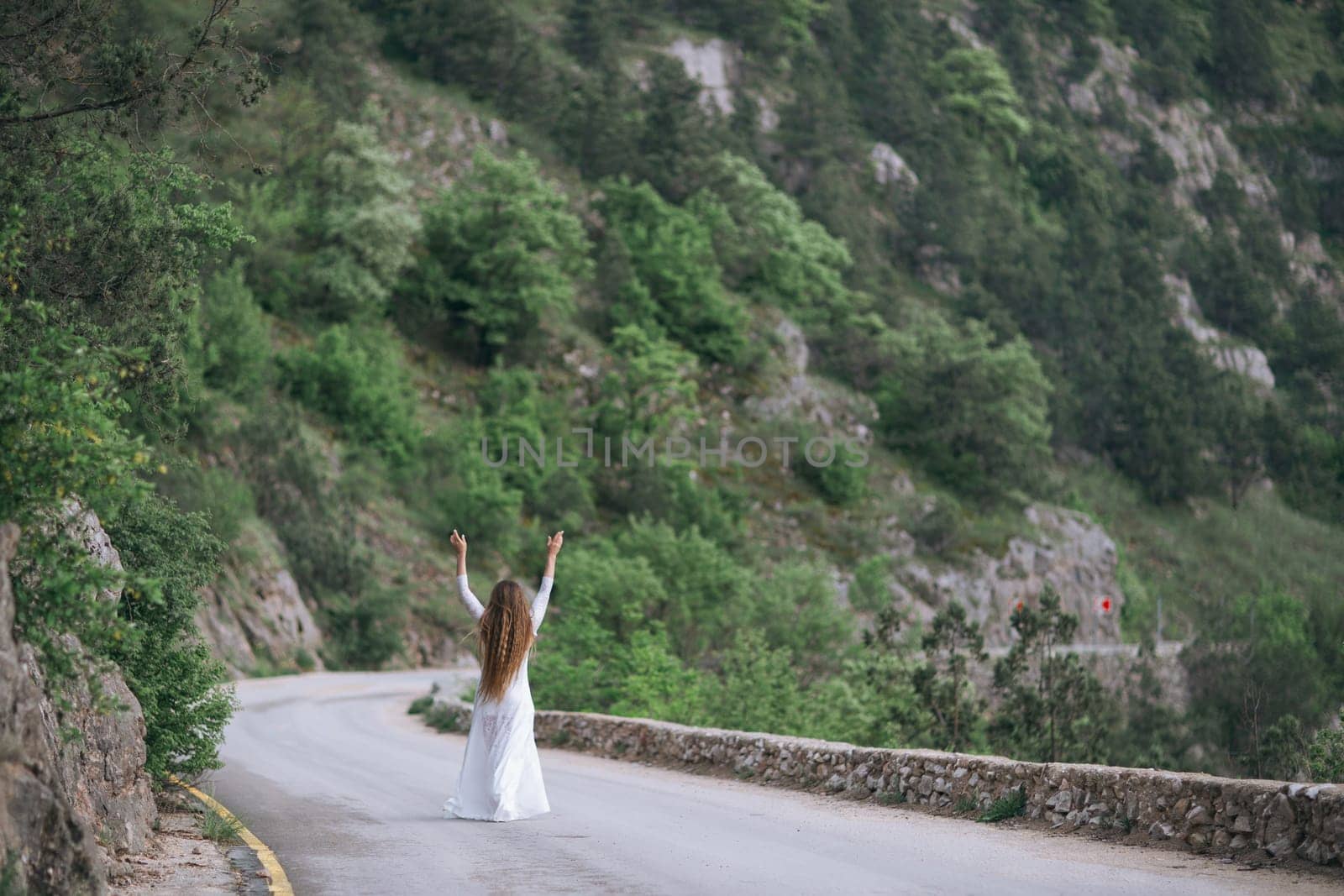 A woman is standing on a road, looking up at the sky. She is wearing a white dress and she is happy. The scene is set in a mountainous area, with trees and rocks in the background