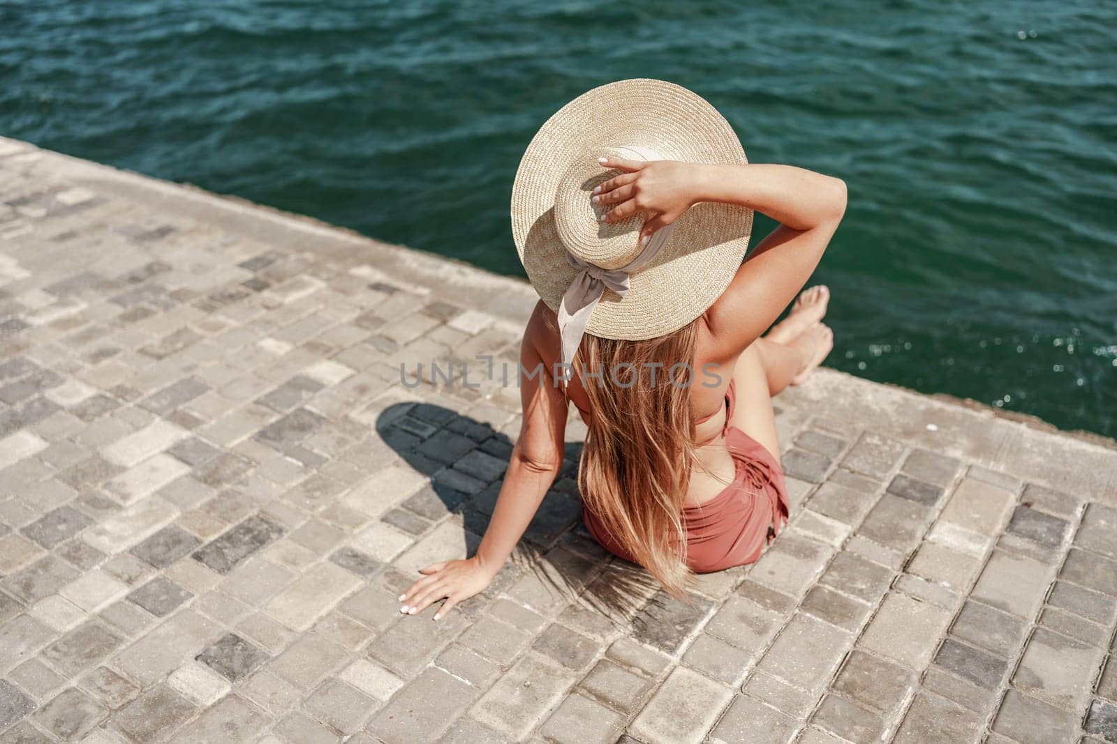 A woman in a swimsuit sits with her back holding a hat, looks at the ocean, sunny day, relaxes