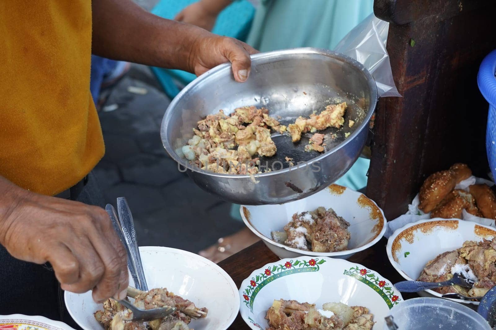 A street food vendor in Surabaya, Indonesia, is preparing a hearty soup with beef ribs and other ingredients. He's using a large metal bowl to mix the ingredients before serving them in smaller bowls.