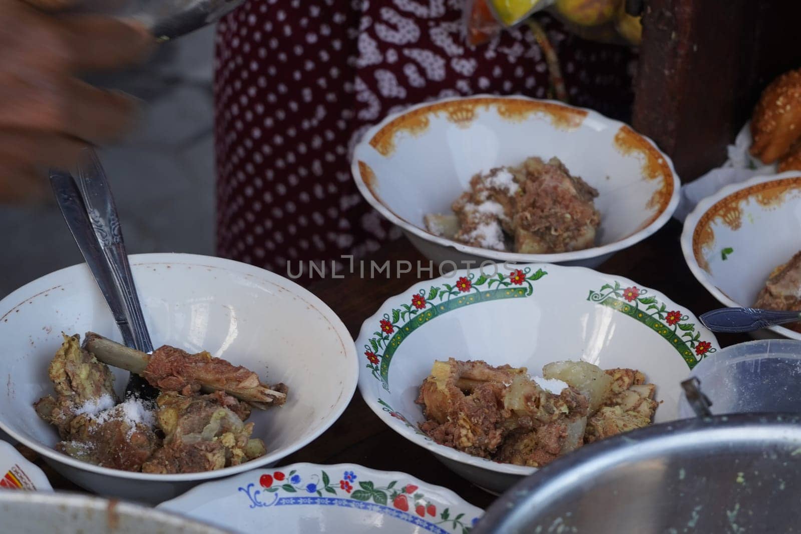 A street food vendor in Surabaya, Indonesia, is adding cooked beef ribs to bowls of soup. The vendor is using tongs to carefully place the ribs into the bowls, which are decorated with colorful patterns.