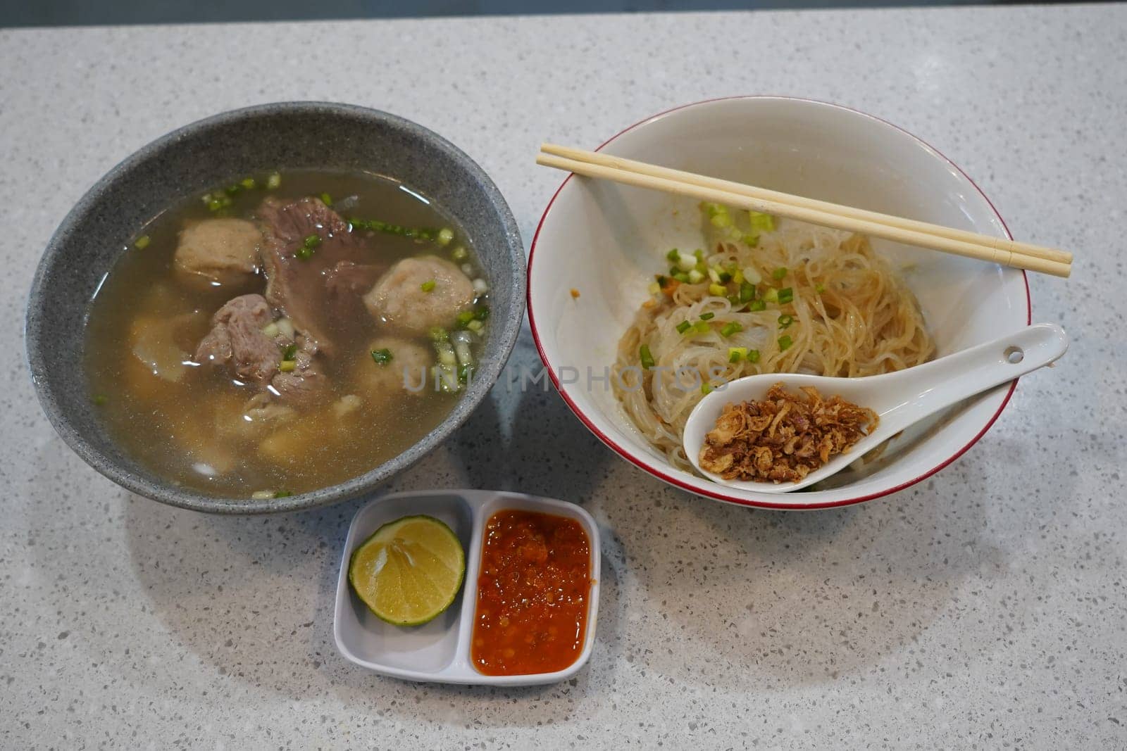 A bowl of savory bakso soup with meatballs, noodles, and broth, served with a small plate of lime wedges and chili sauce, accompanied by a bowl of vermicelli noodles and a small plate of fried shallots.