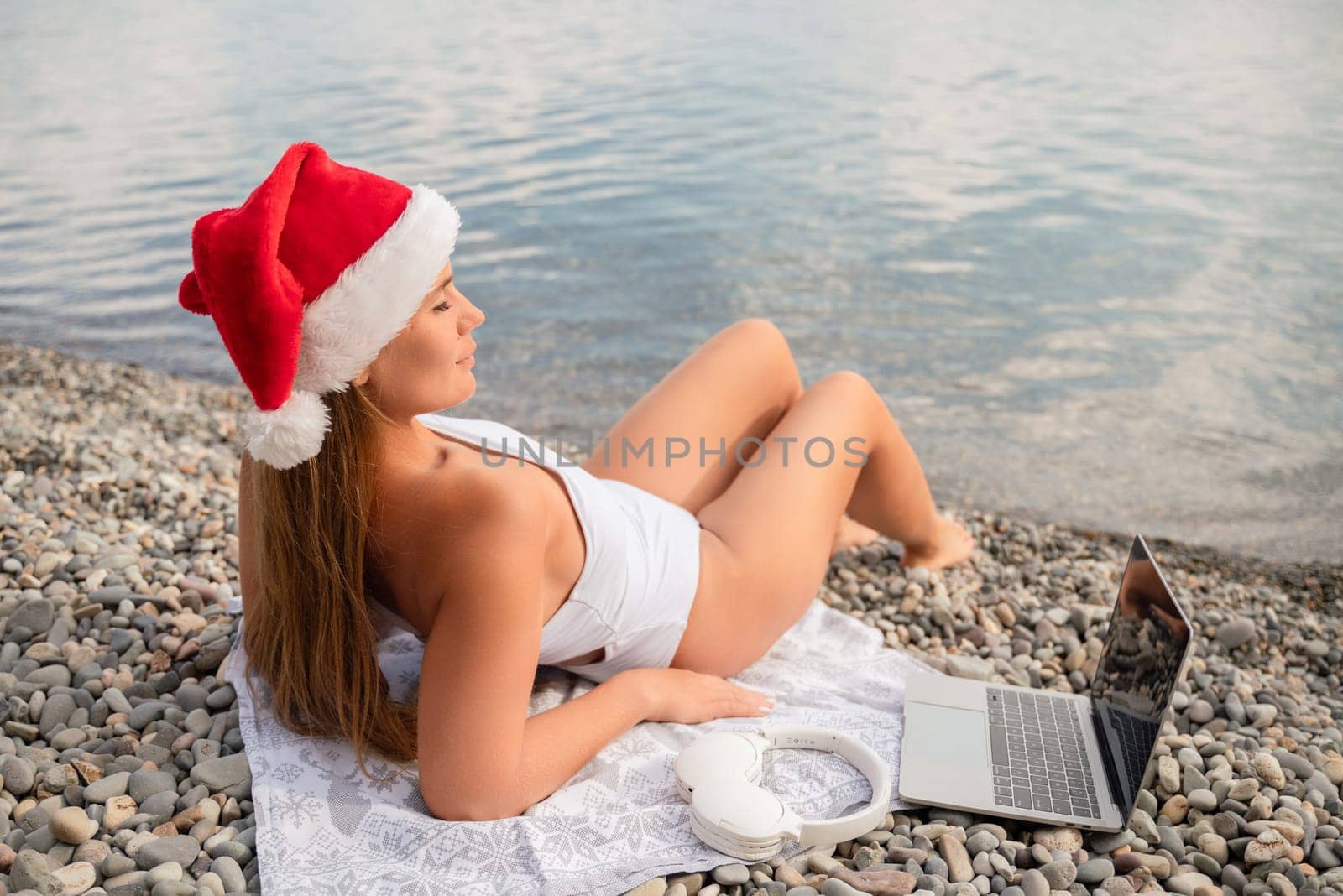 A woman is laying on the beach with a laptop and headphones on her head. She is wearing a red Santa hat