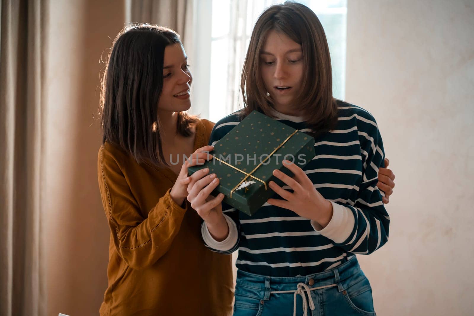 Two happy young girls laugh, friends hug and give each other gifts during the holiday. Women surprise and congratulate on the anniversary of their relationship in their cozy home.