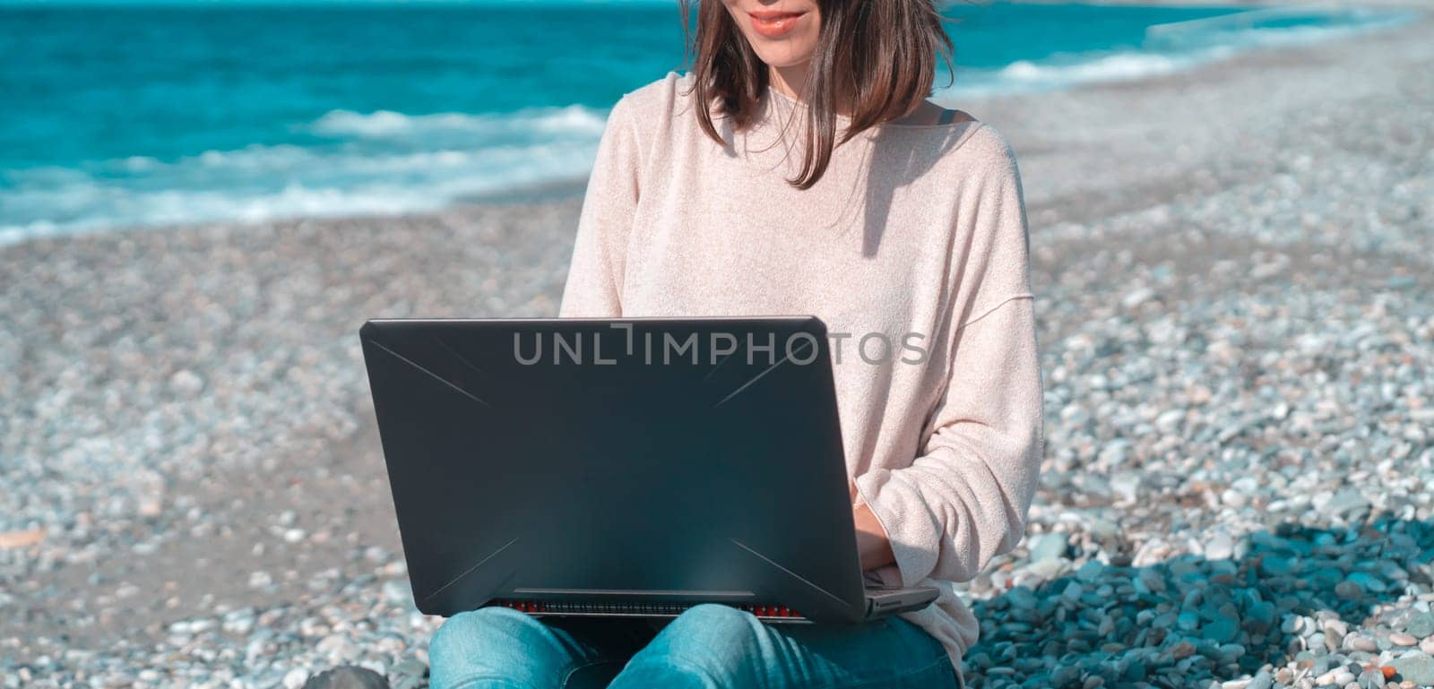 A young smiley girl in a hat and casual sweater sits on the beach by the sea with a laptop on a sunny day, works, studies, buys tickets during trip, woman rests on vacation and types on the keyboard.