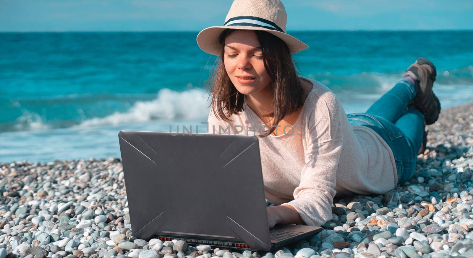 A young girl in a light hat and casual sweater lies on the beach by the sea with a laptop on a sunny day, works, studies, buys tickets during trip, a woman rests on vacation and types on the keyboard.