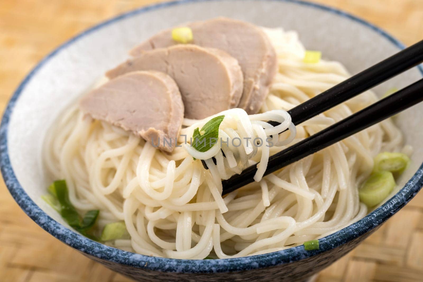 A close up photo of using chopsticks  to eat a bowl of noodles topped with pork slices and green onions.