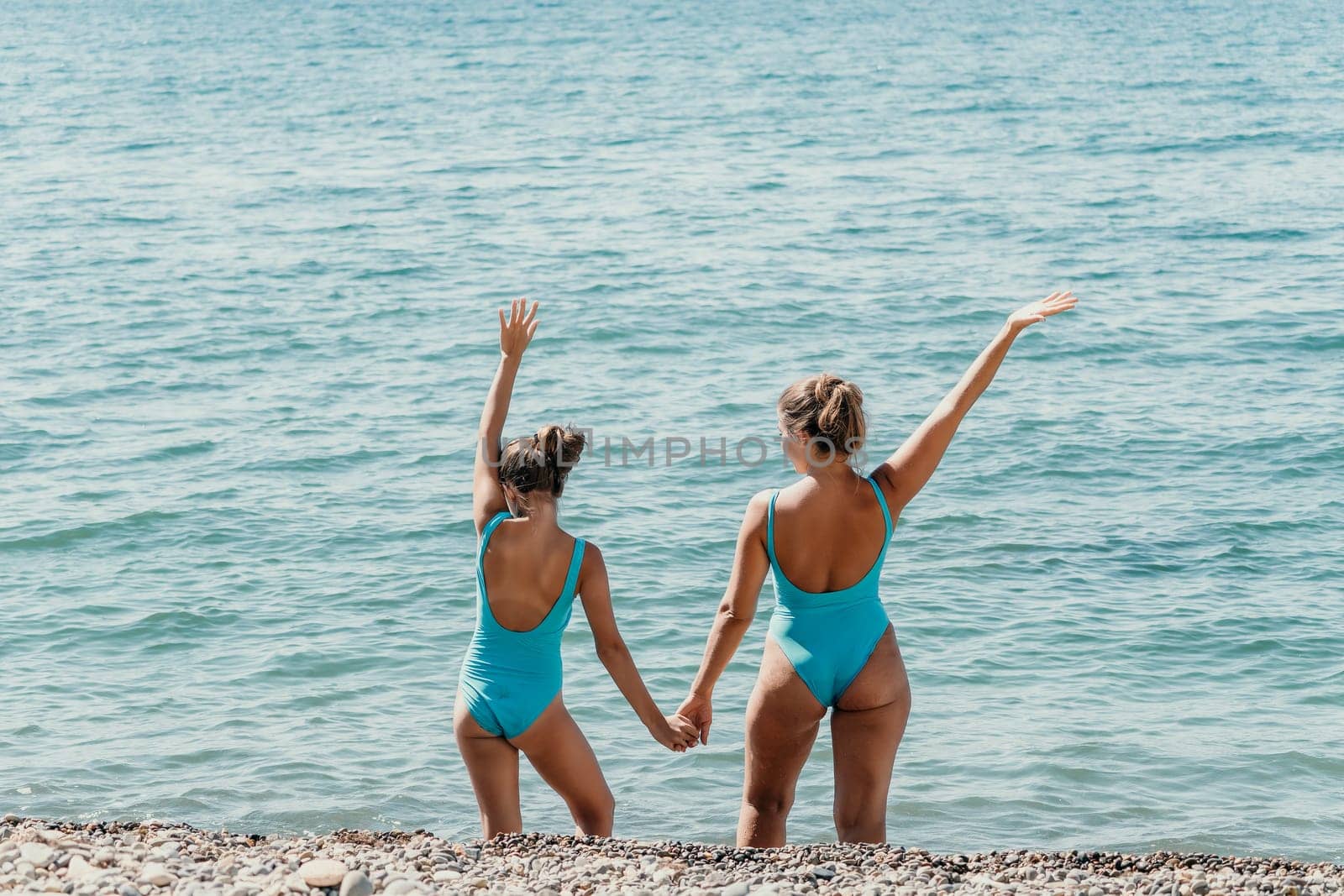 A mother and her daughter stand hand in hand on a pebble beach, looking out at the sparkling blue sea.