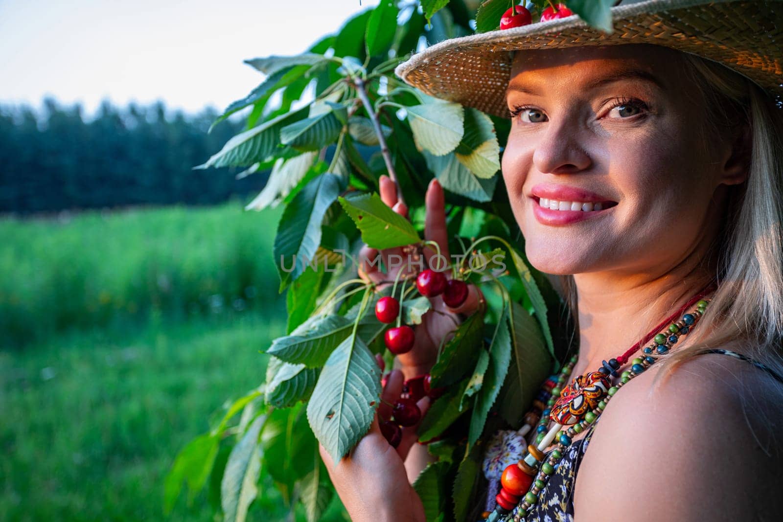 In the serene ambiance of a countryside landscape, a vision of beauty emerges. A radiant woman, her blonde hair framing her face, gazes upwards towards the heavens and the setting sun. The golden rays of sunlight gracefully illuminate her features, accentuating the emerald green of her eyes and the warmth of her smile, revealing her pearly white teeth. Clad in light summer attire, she exudes an air of casual elegance, her ensemble complemented by a colorful beaded necklace adorning her neck and a straw hat atop her head. One hand delicately cradles a branch of the cherry tree. The woman is looking into the lens. Behind her, the cherry tree stands tall, its branches reaching towards the azure sky, while the rural landscape stretches out beyond, a picturesque backdrop to the tranquil moment captured in the photograph. In this idyllic setting, amidst the beauty of nature, the woman finds serenity and joy, a harmonious blend of elegance and rustic charm in the simplicity of the countryside.