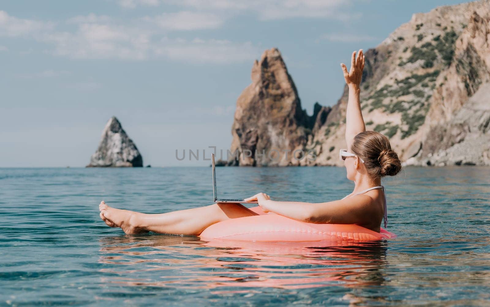 Woman freelancer works on laptop swimming in sea on pink inflatable ring. Pretty lady typing on computer while floating in the sea on inflatable donut at sunset. Freelance, remote work on vacation