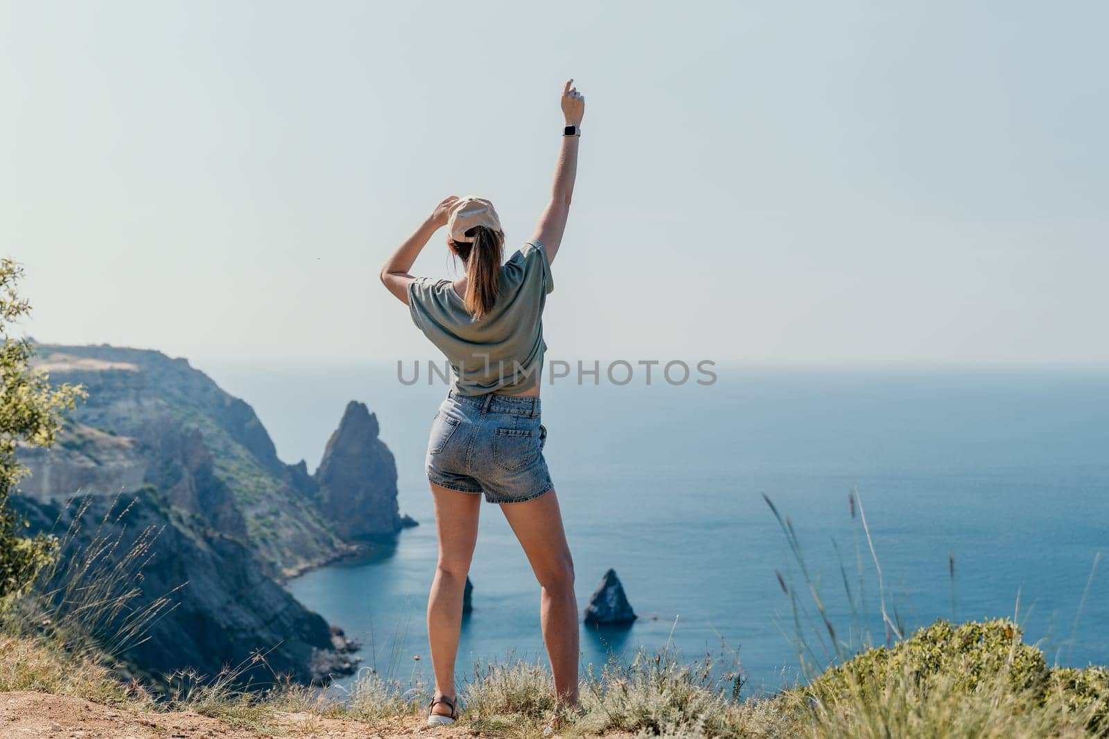 A young woman in a green shirt and denim shorts stands on a cliff overlooking a picturesque coastline.